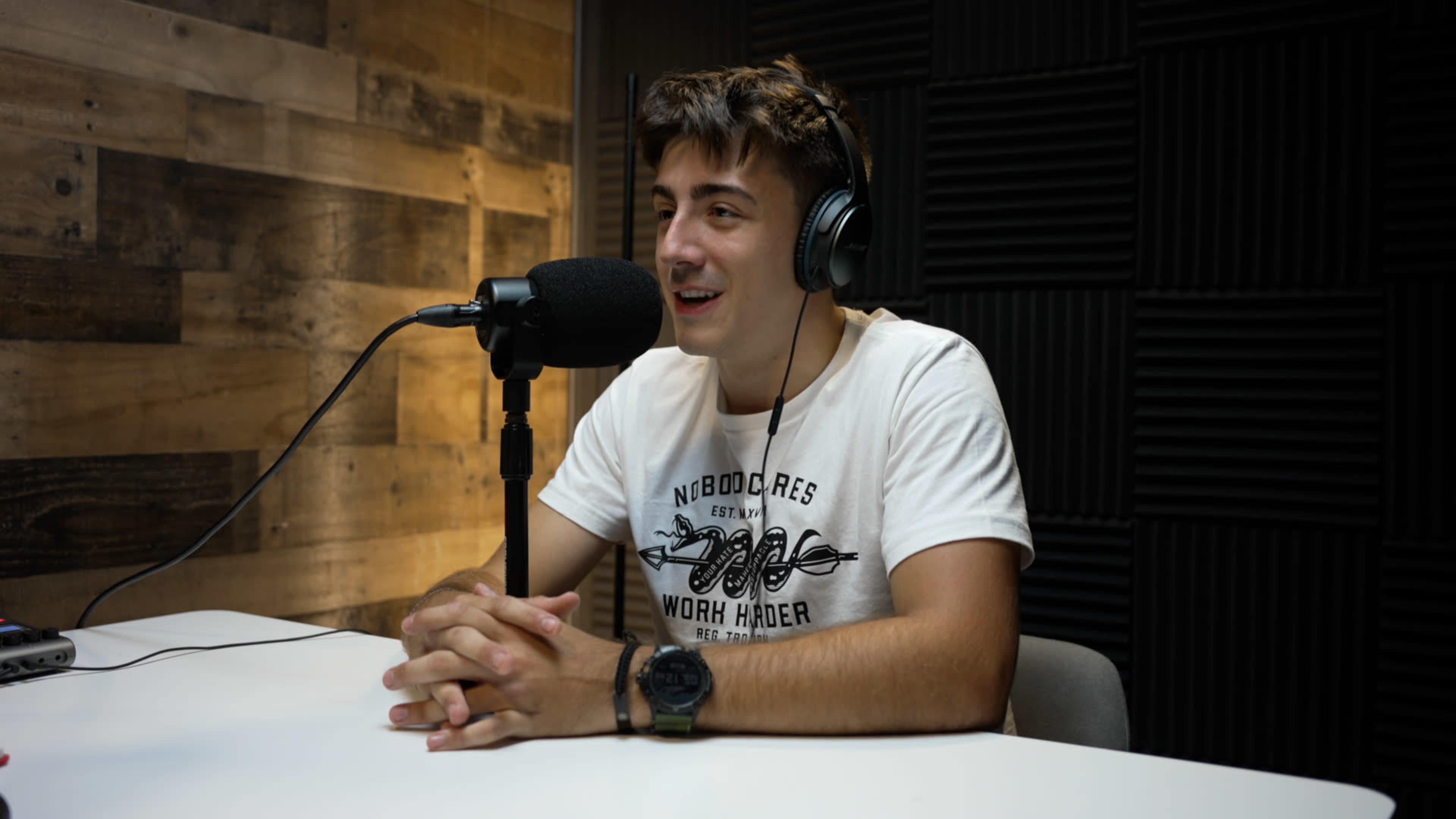 A young man wearing headphones sits at a table in a recording studio with a microphone in front of him.