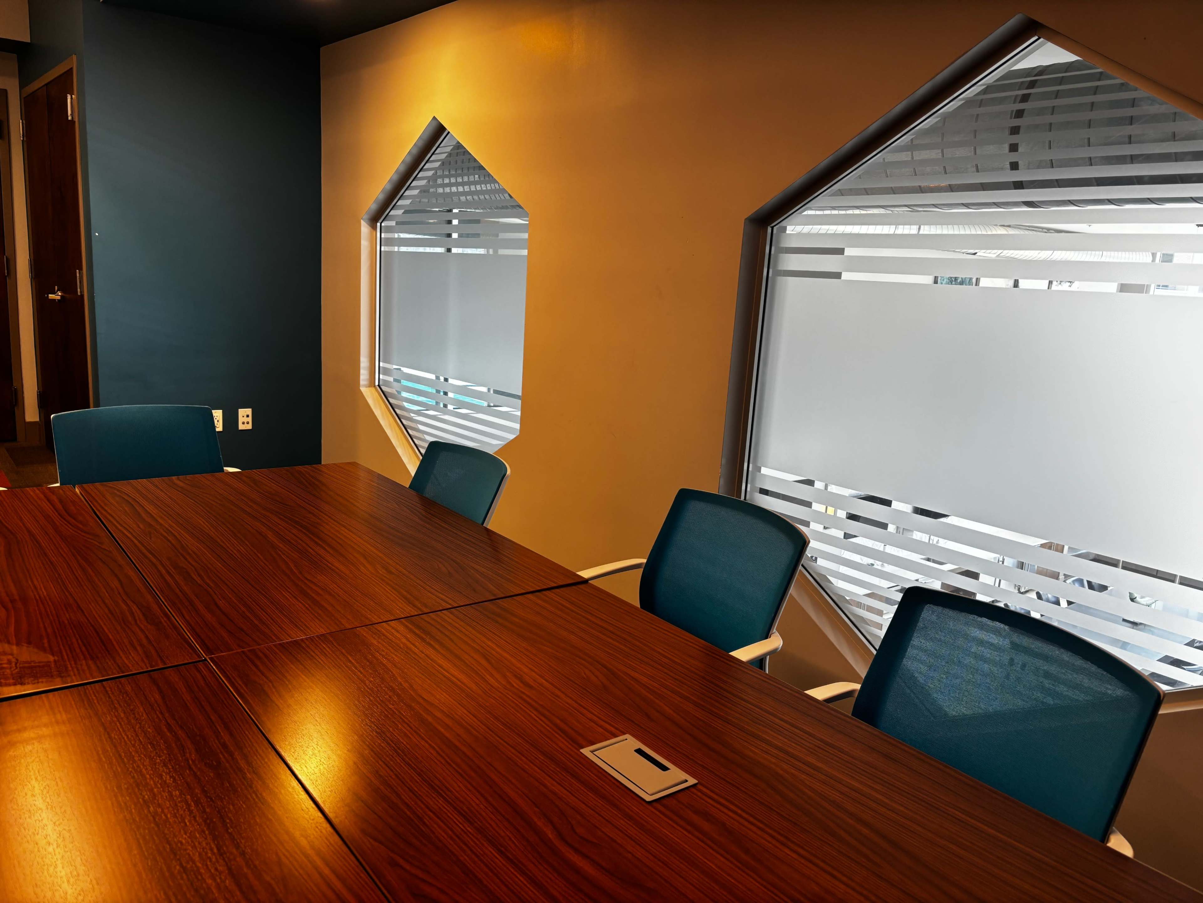 A modern conference room features a wooden table surrounded by blue chairs, with hexagonal windows allowing light to enter.