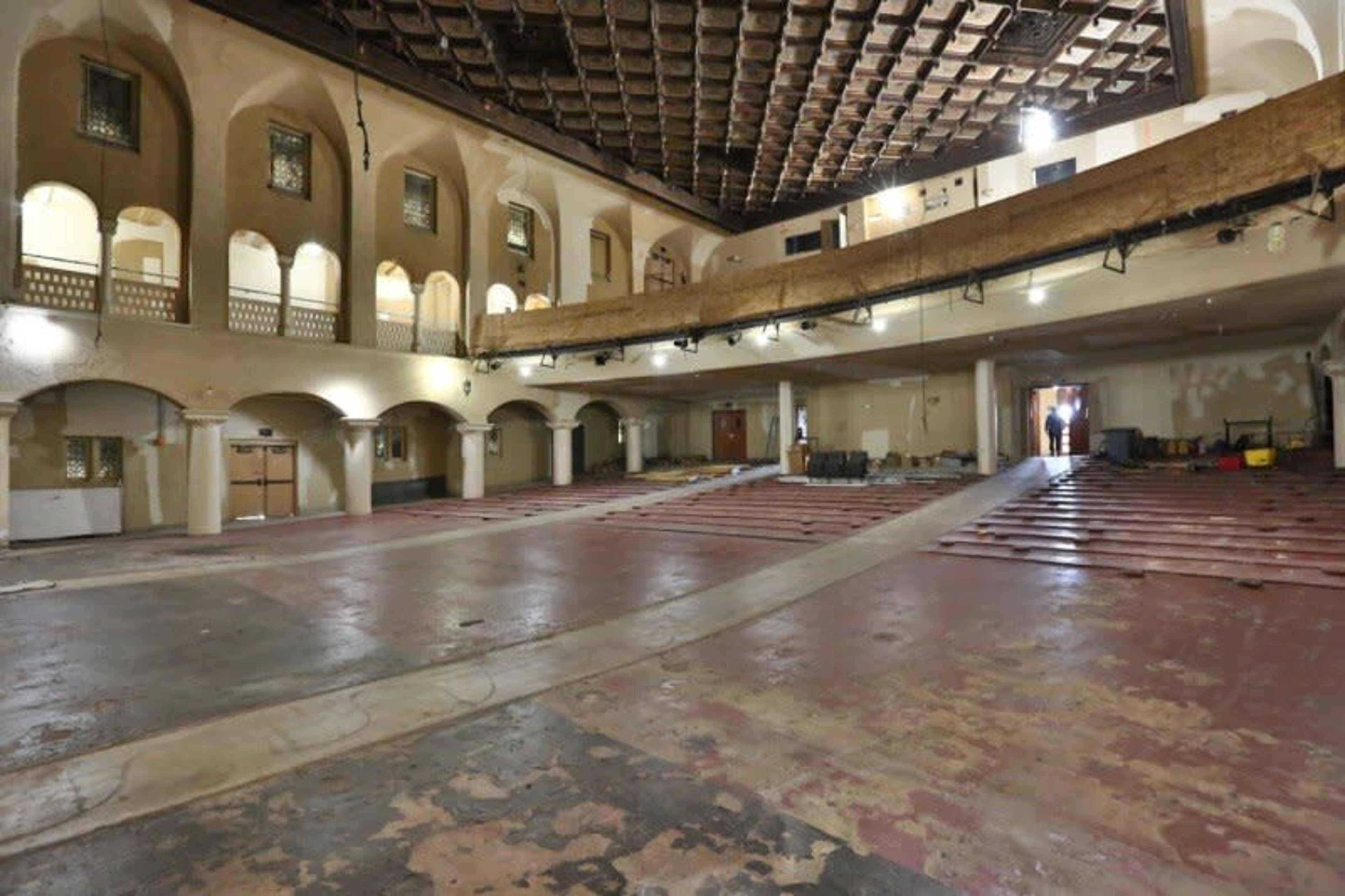 A large, empty indoor space with a wooden balcony overlooking a faded parquet floor and walls adorned with arched windows.