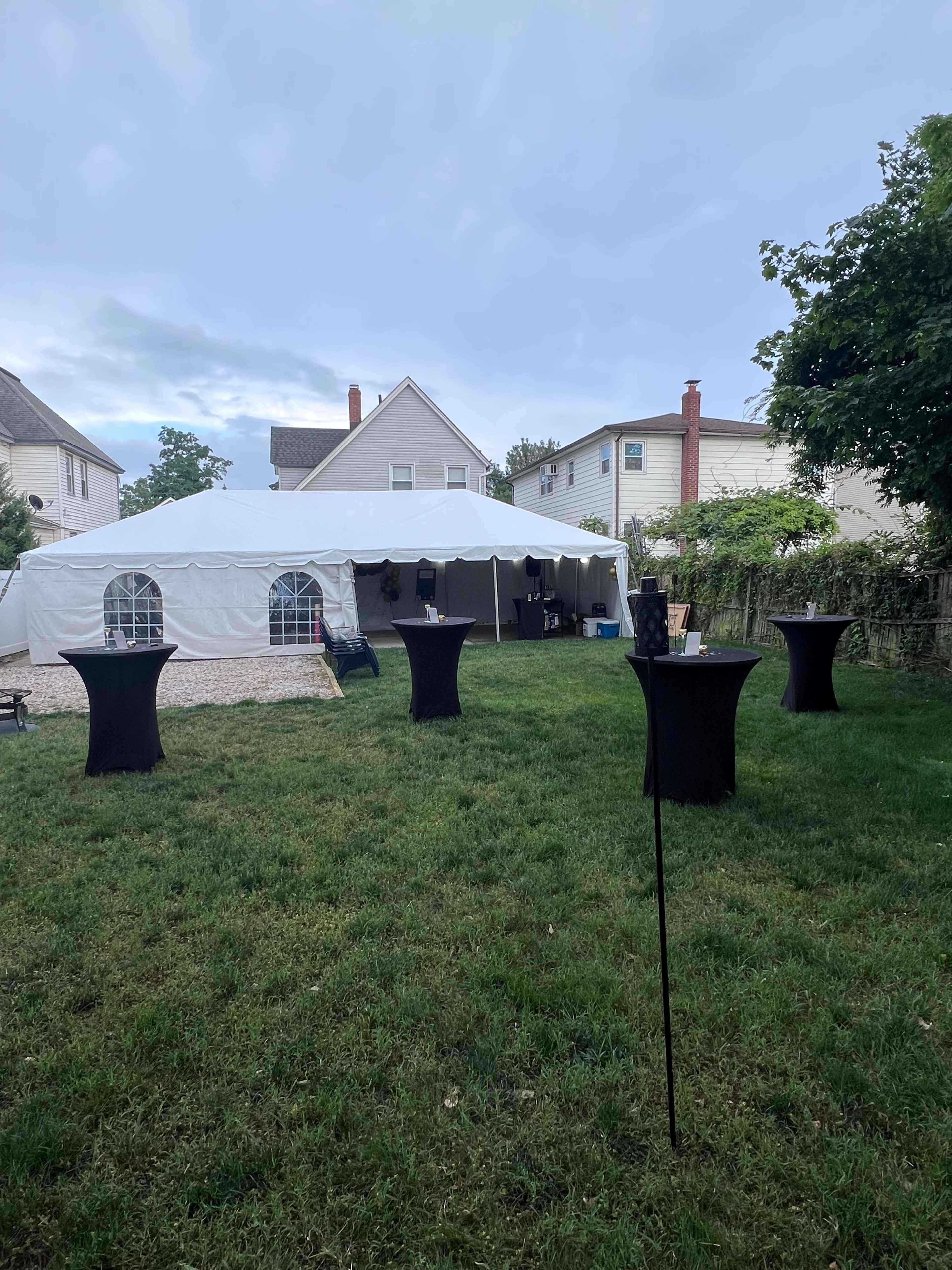 A white canopy tent is set up in a grassy backyard, surrounded by tall tables.