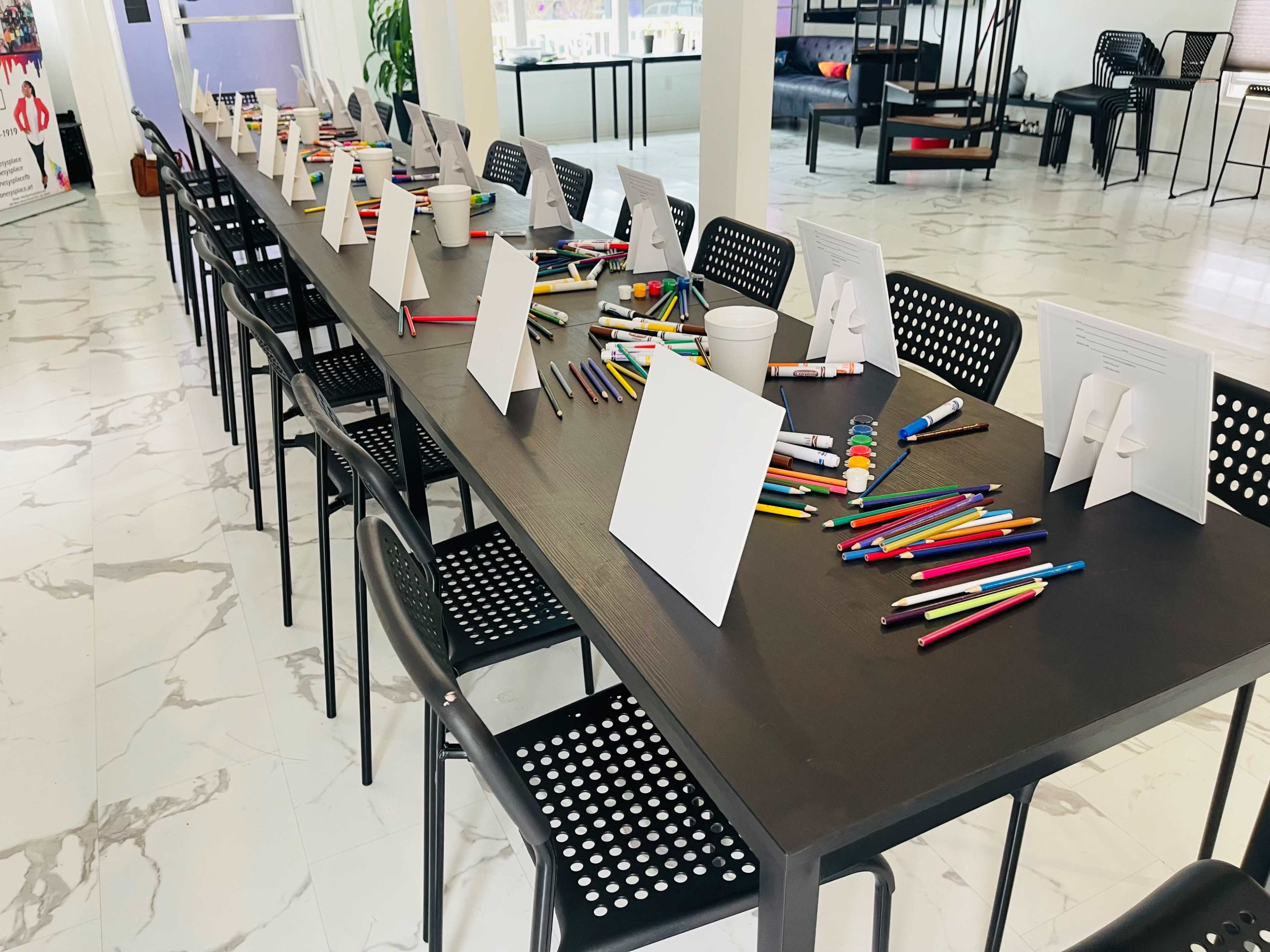 A long table is set up with various colored pens and blank cards, ready for an activity, surrounded by black chairs in a brightly lit room.