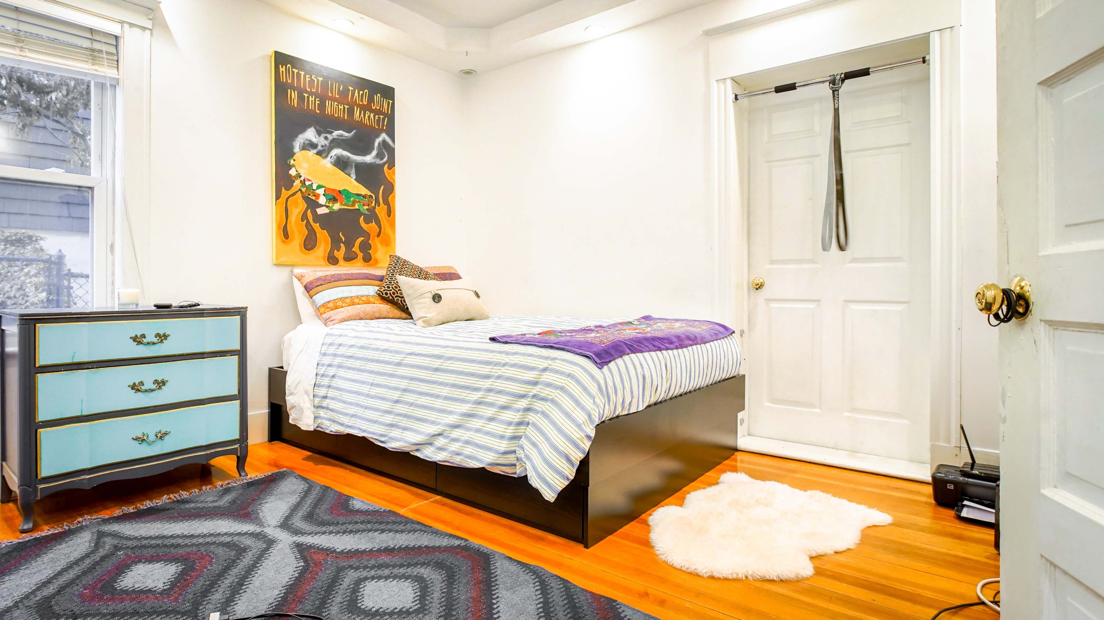 A bedroom features a bed with striped bedding, a colorful artwork on the wall, a blue dresser, and a small area rug.