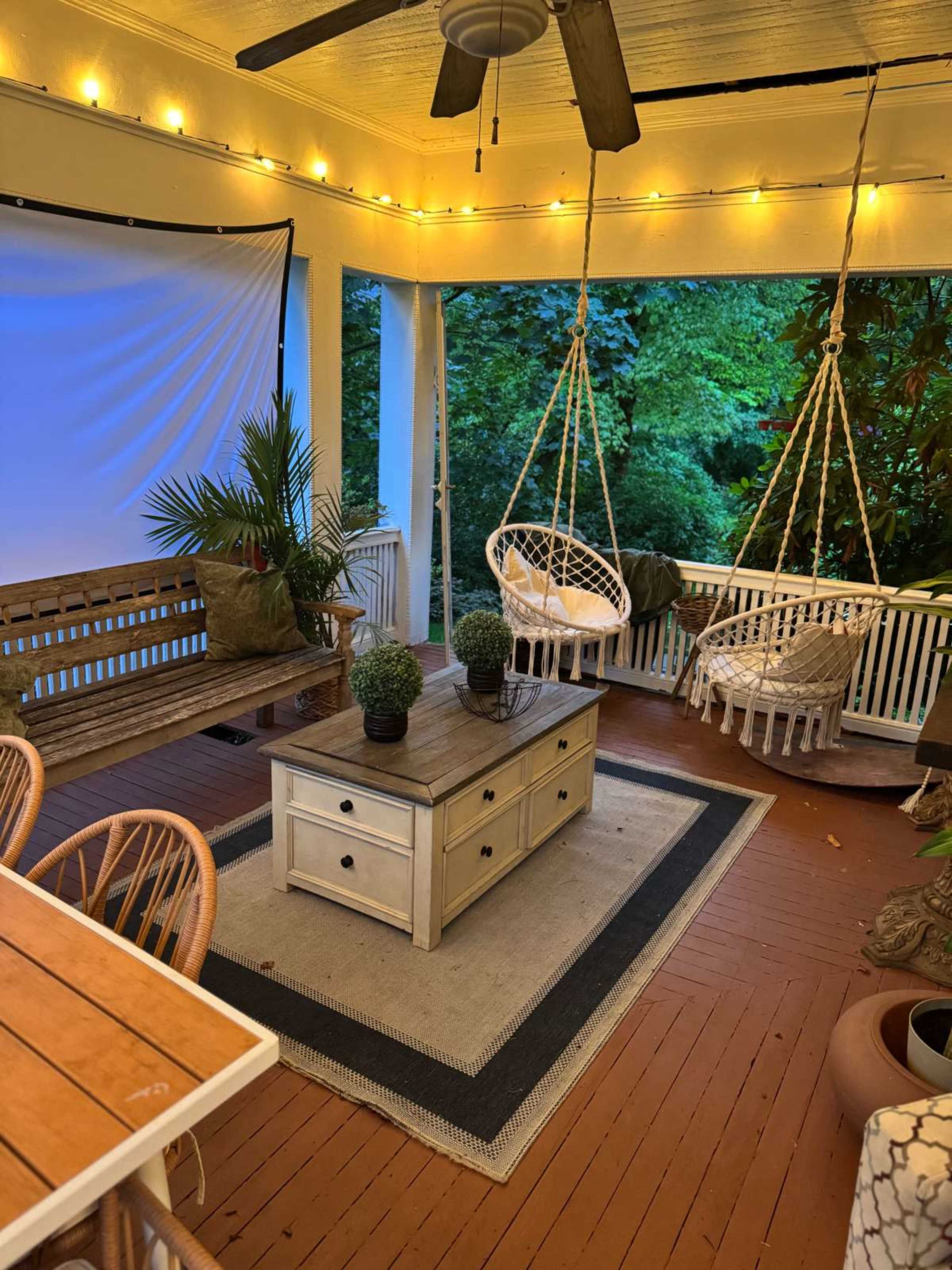 The image shows a cozy porch with hanging chairs, a wooden coffee table, and decorative plants, surrounded by string lights and greenery.