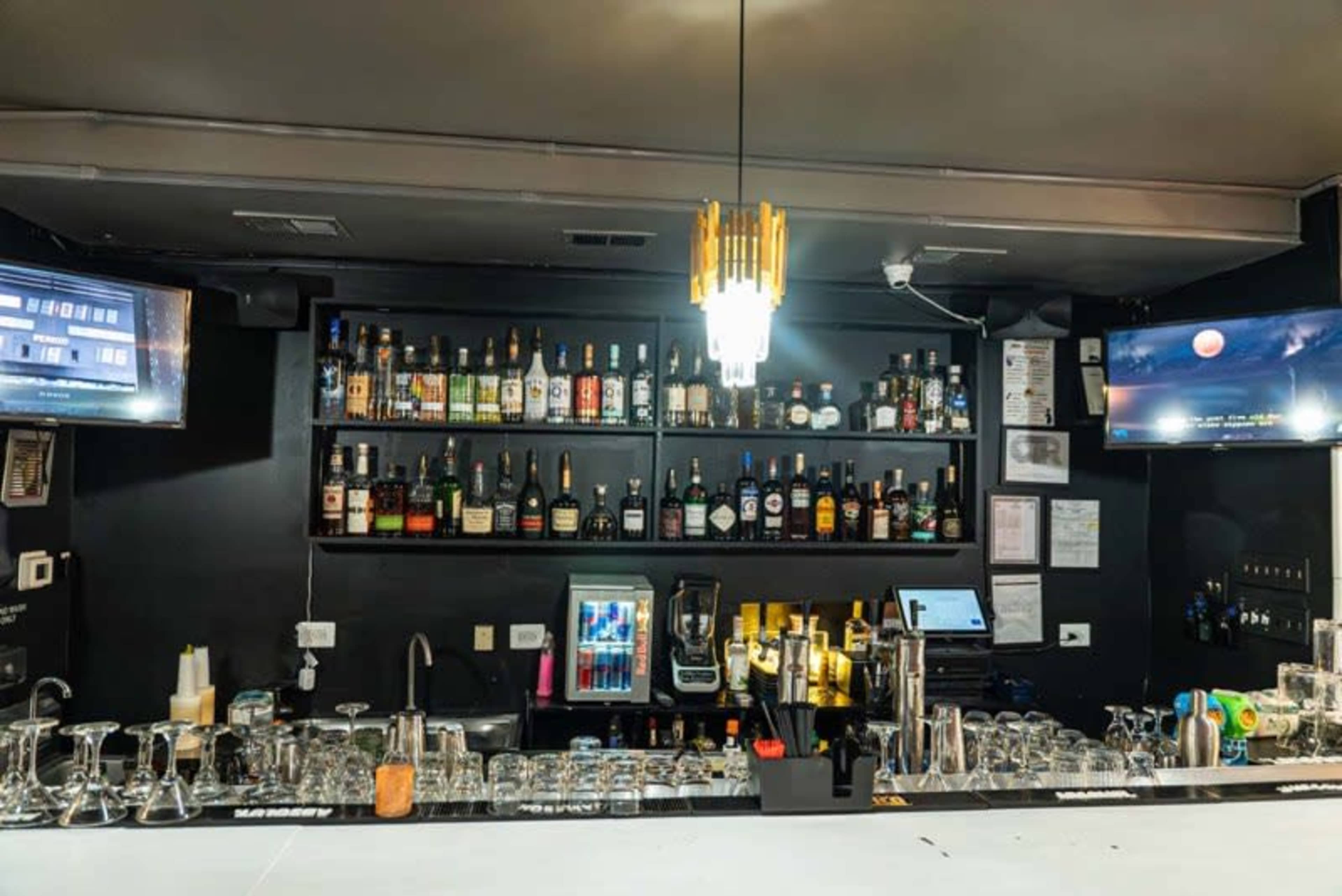 The image shows a bar featuring shelves stocked with various bottles of liquor, a cash register, and multiple screens displaying content.