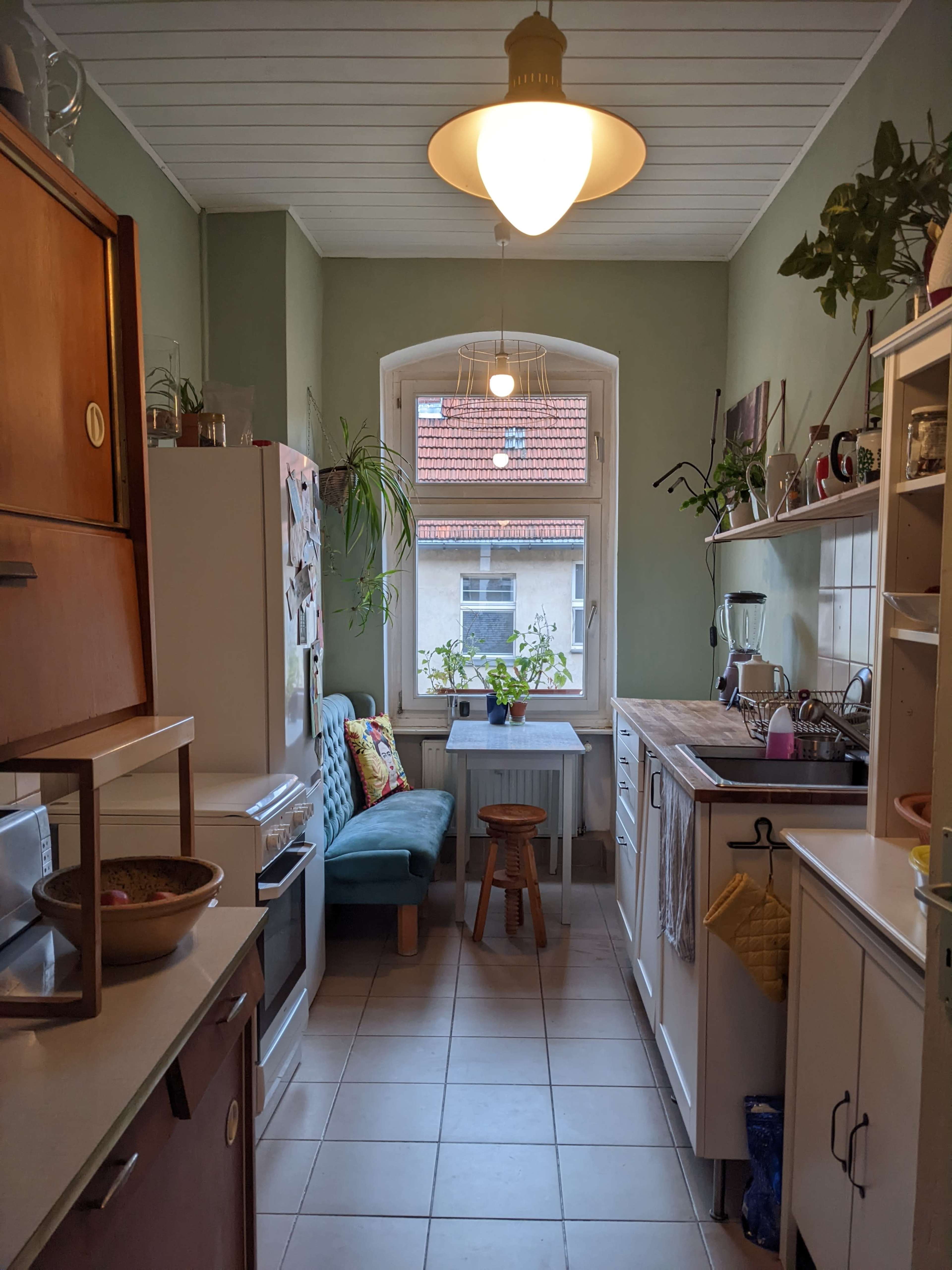 The kitchen features a long layout with a window seat, wooden countertops, a sink, and various plants along the window sill.