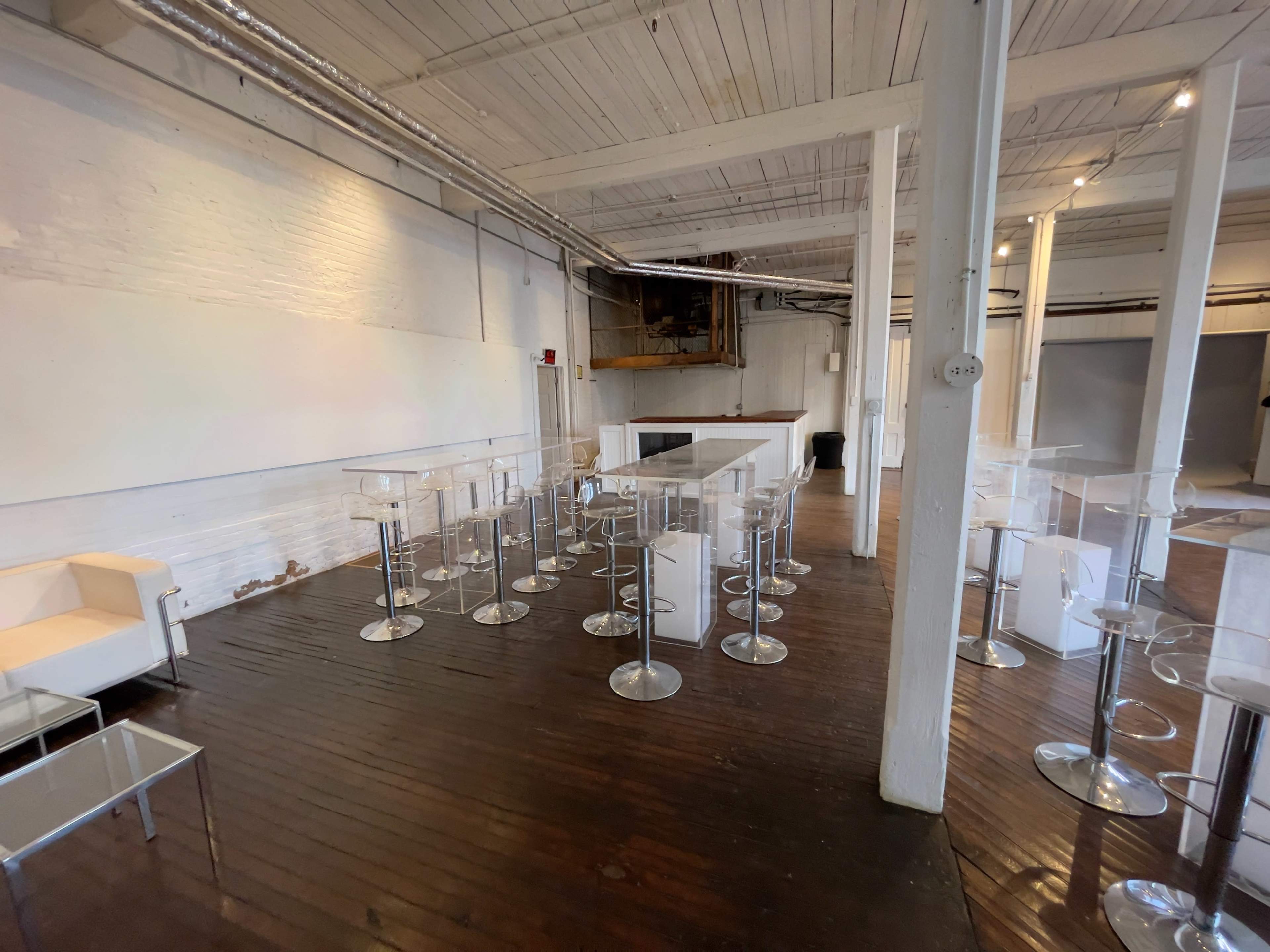 An empty event space with clear high-top tables and a few white seating areas, set against a wooden floor and exposed beams.