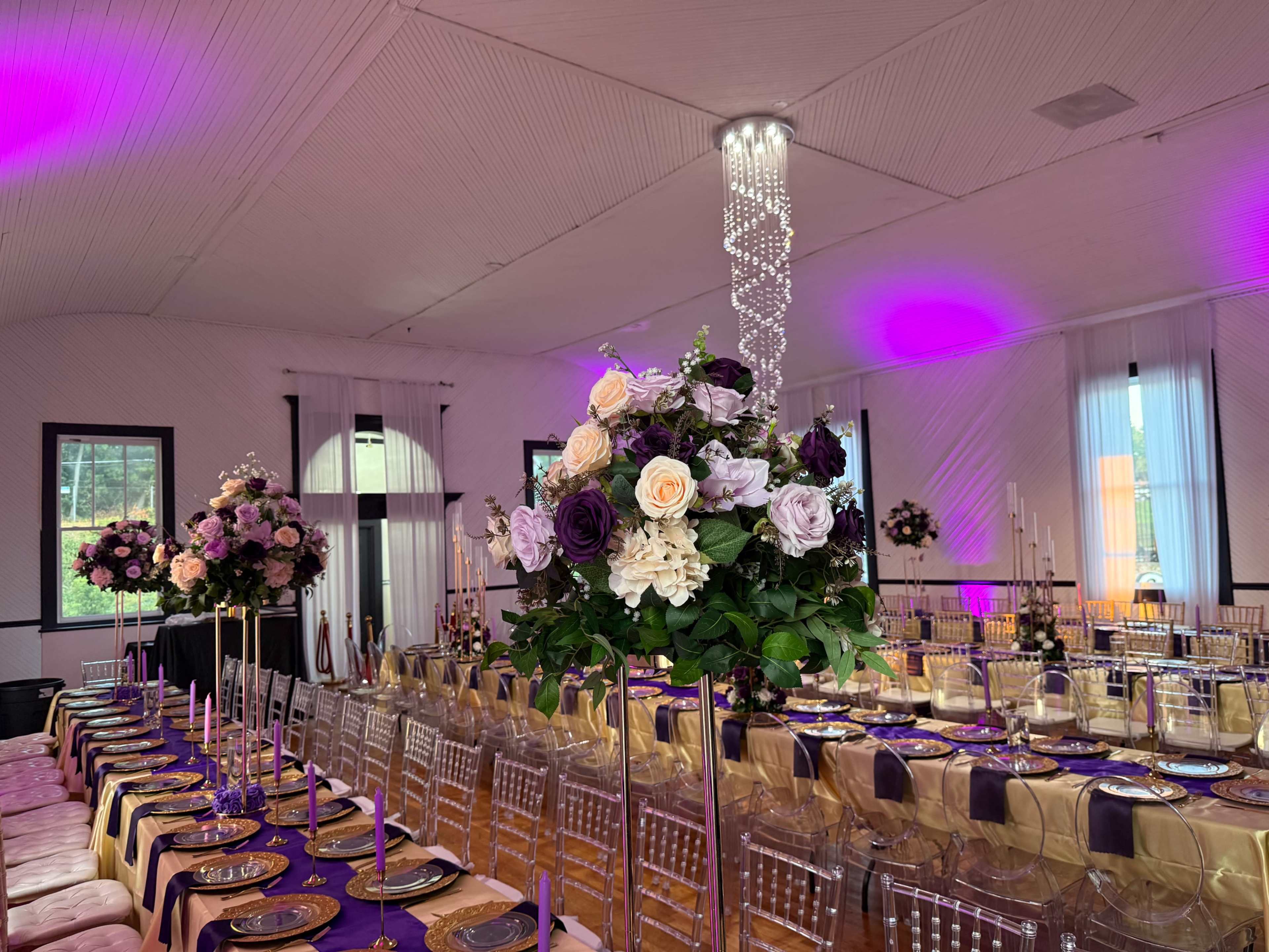 The image shows a banquet hall set for an event, featuring elegantly arranged tables adorned with floral centerpieces and decorative lighting.