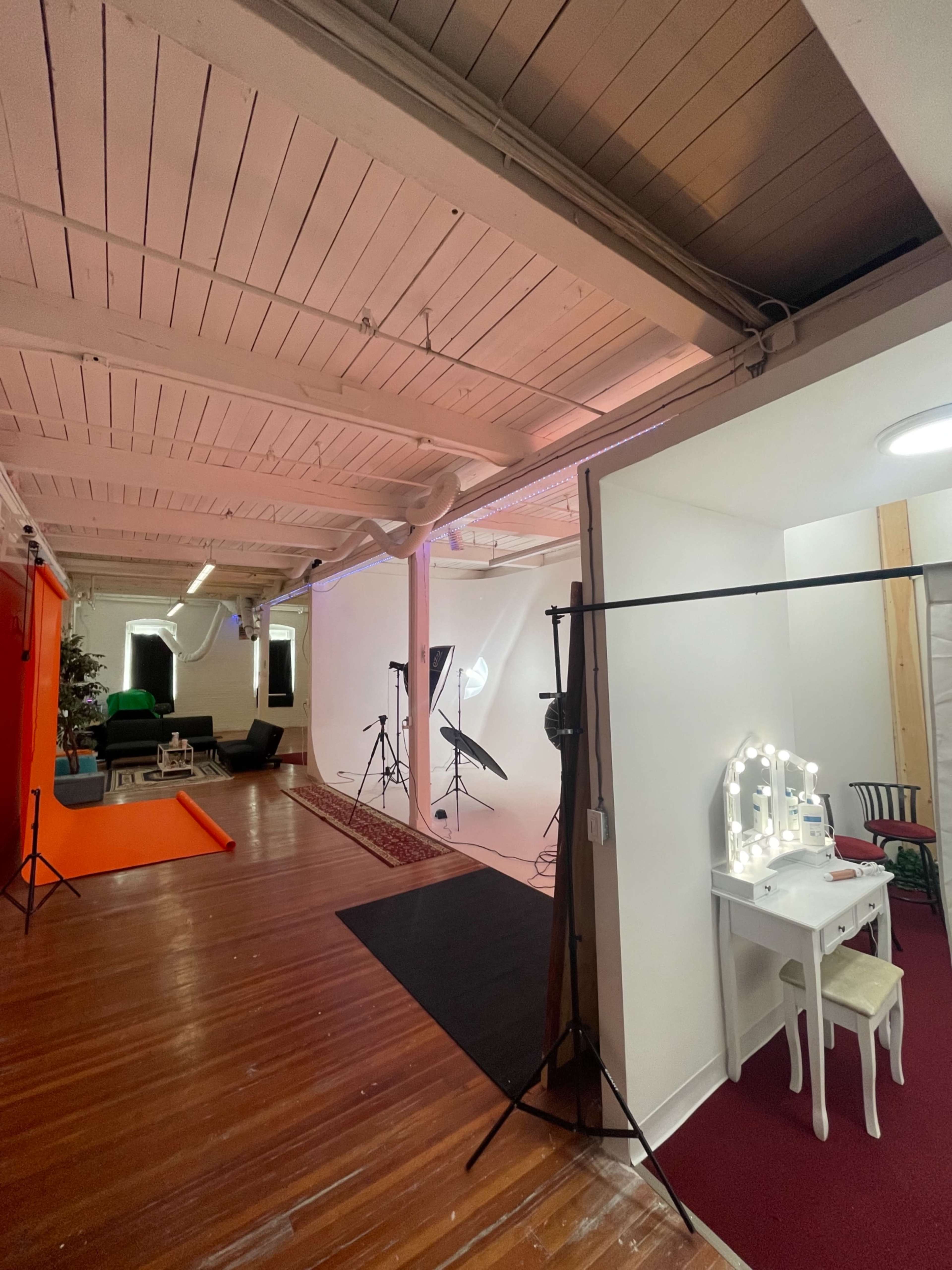 The image shows a spacious studio with a vibrant orange backdrop on one side and a well-lit area featuring photography equipment on the other, along with a vanity table and chair.