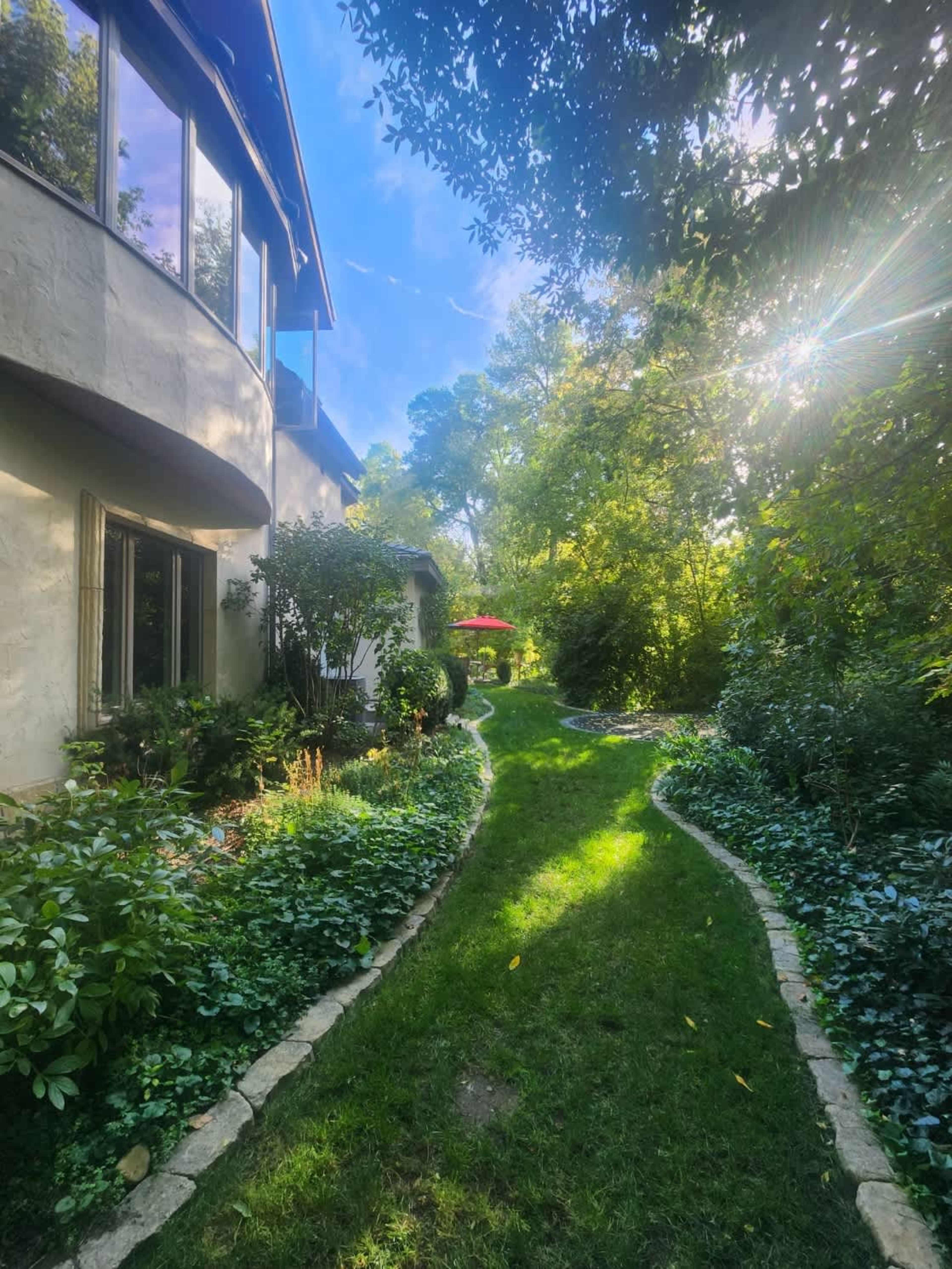 A winding path cuts through a lush garden, leading toward a red umbrella in the distance under a canopy of trees.