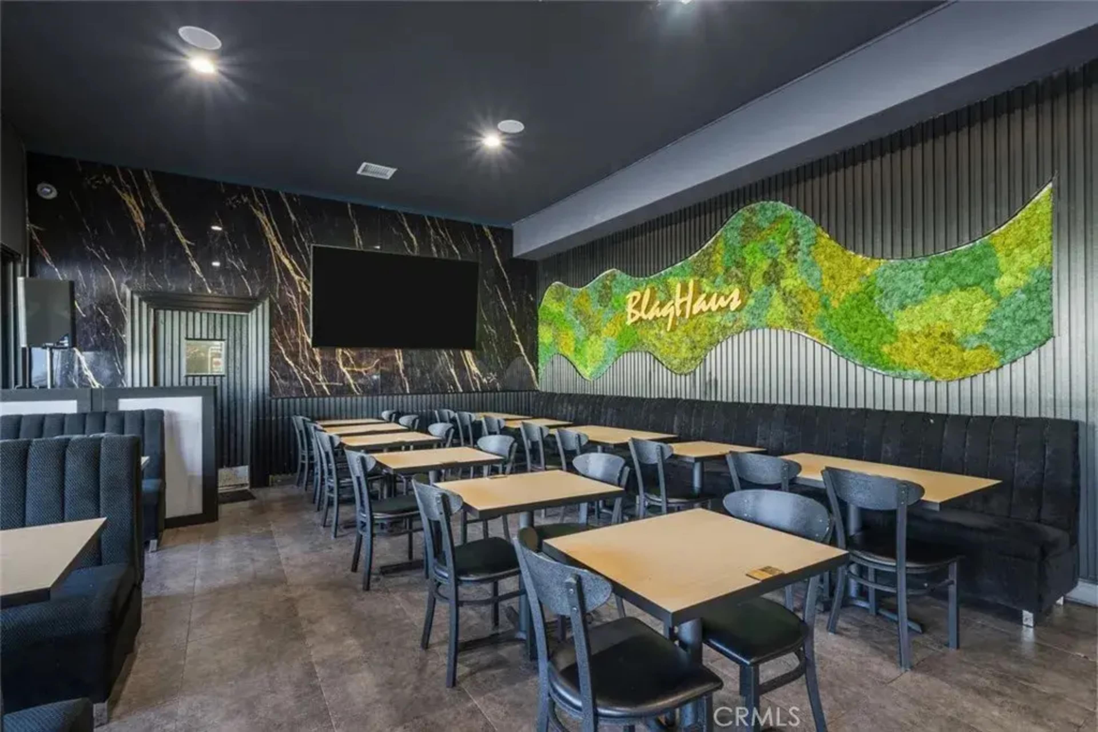 The image shows a modern restaurant interior featuring black seating, wooden tables, and a decorative green wall art piece.