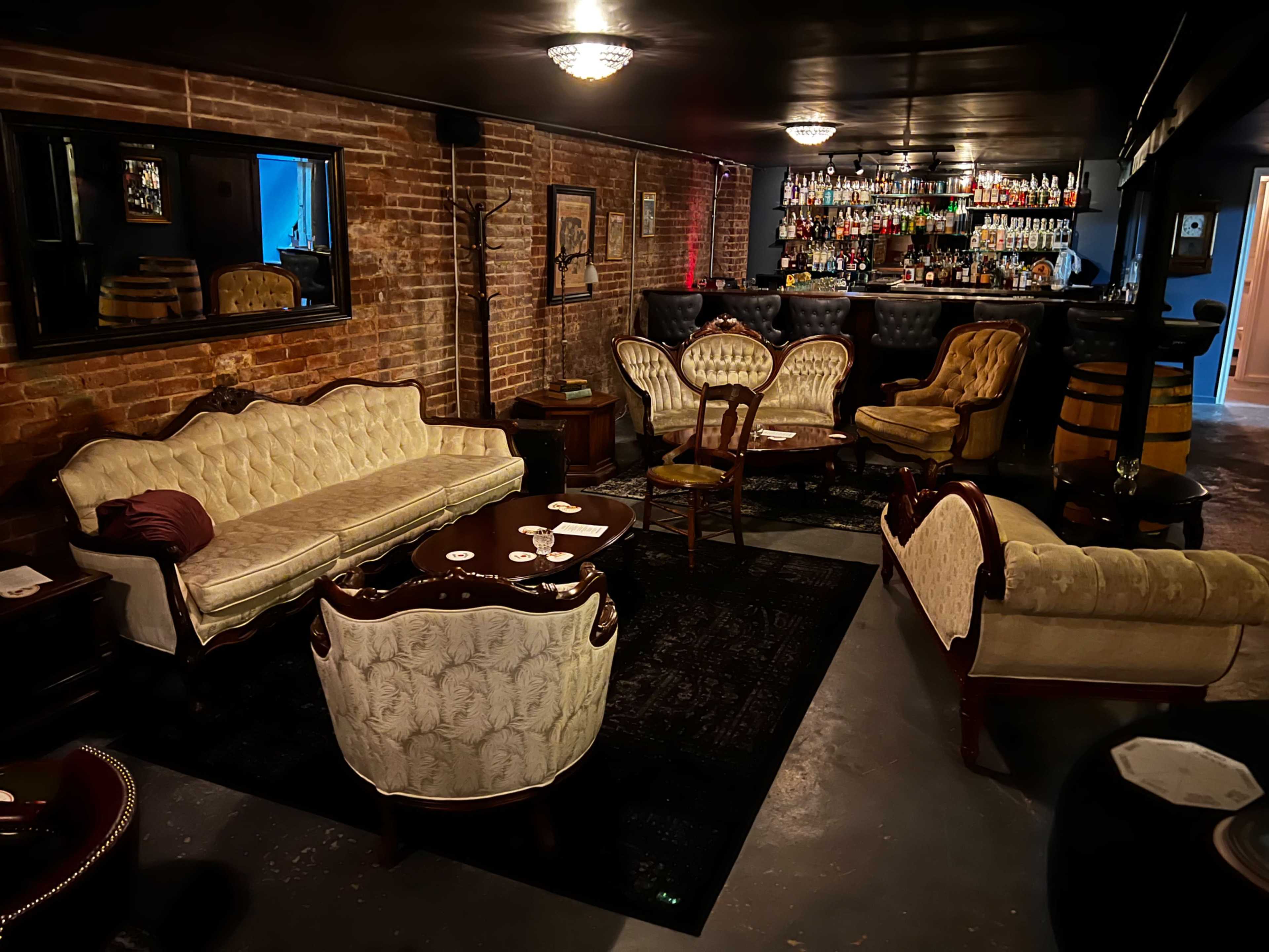 A dimly lit lounge with vintage-style furniture, including ornate sofas and chairs, arranged around a round table in front of a bar lined with liquor bottles.