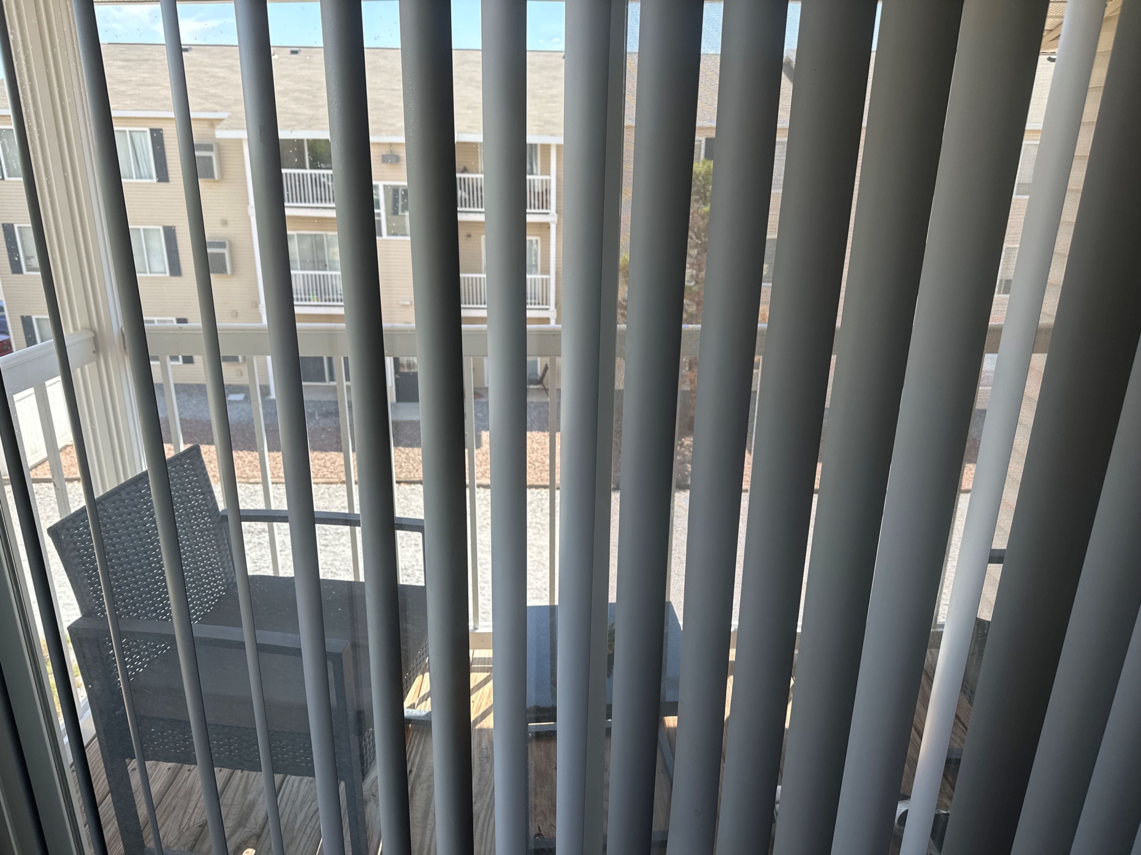 The image shows a view from behind vertical blinds, looking out onto a patio area with outdoor furniture and a gravel surface.