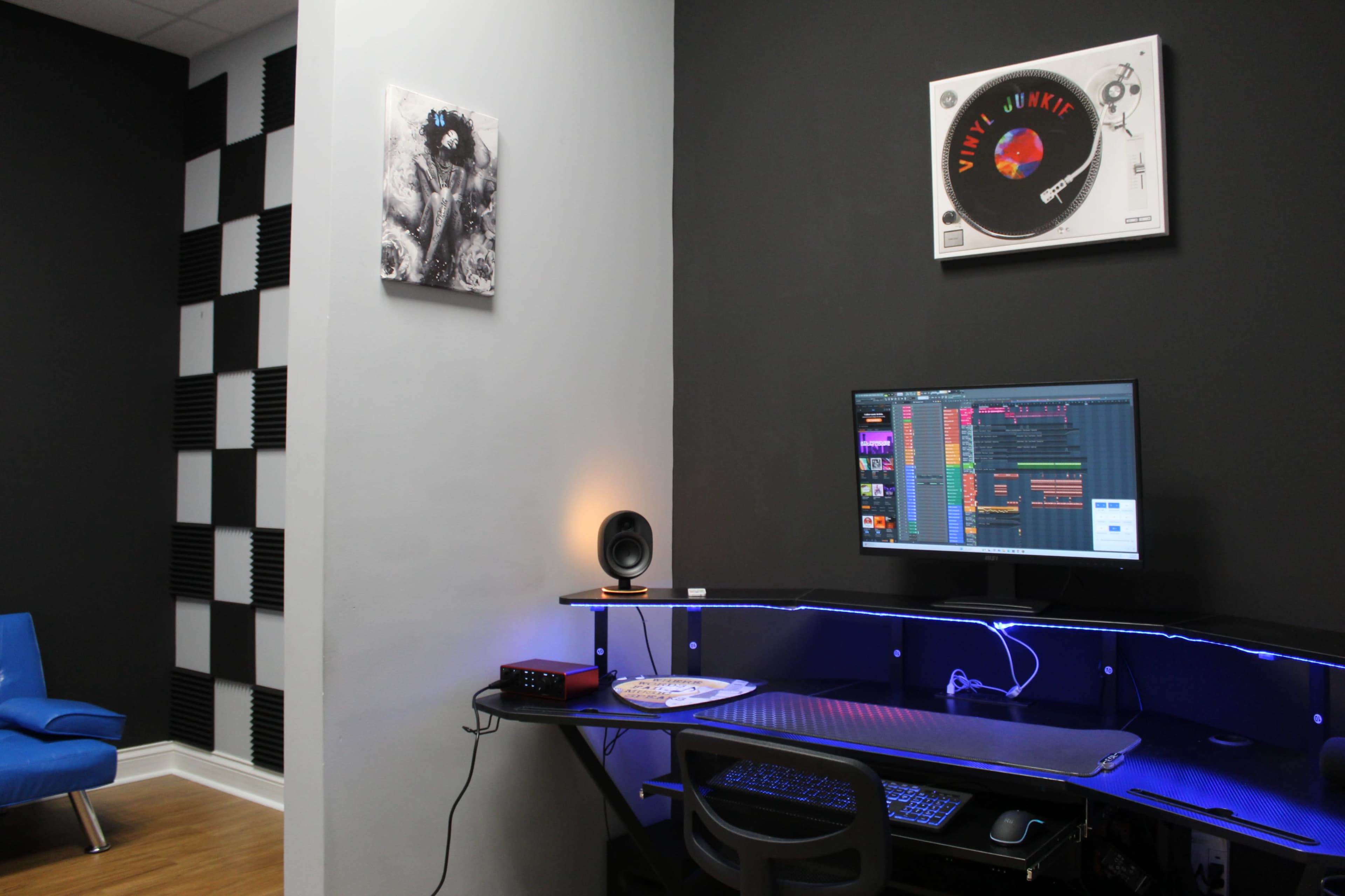 The image shows a modern music production setup featuring a desk with a computer monitor, audio interface, and a speaker, set against a black and white wall design with soundproofing panels.