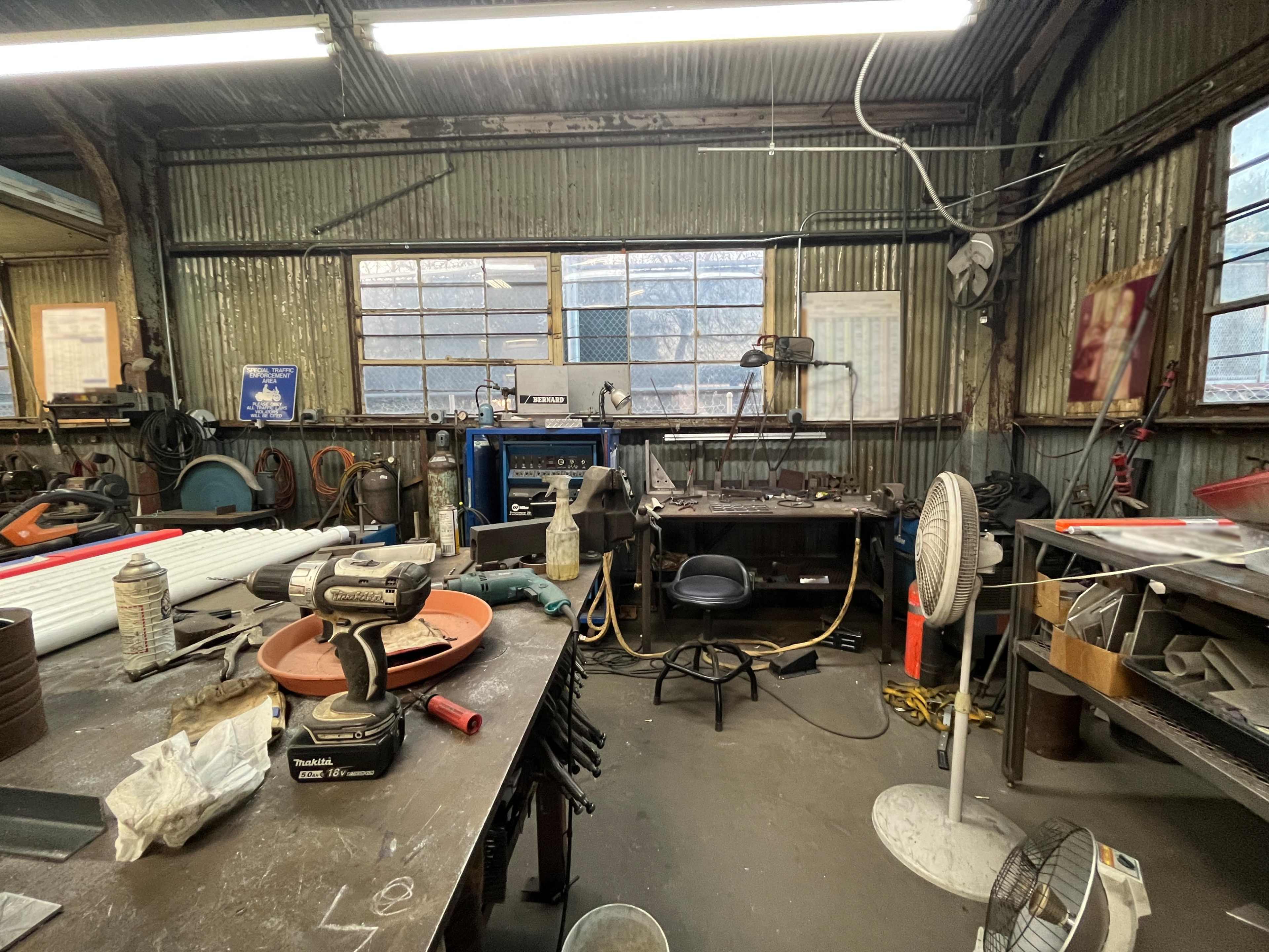 A cluttered workshop filled with tools, equipment, and a workbench, featuring a chair and a standing fan.