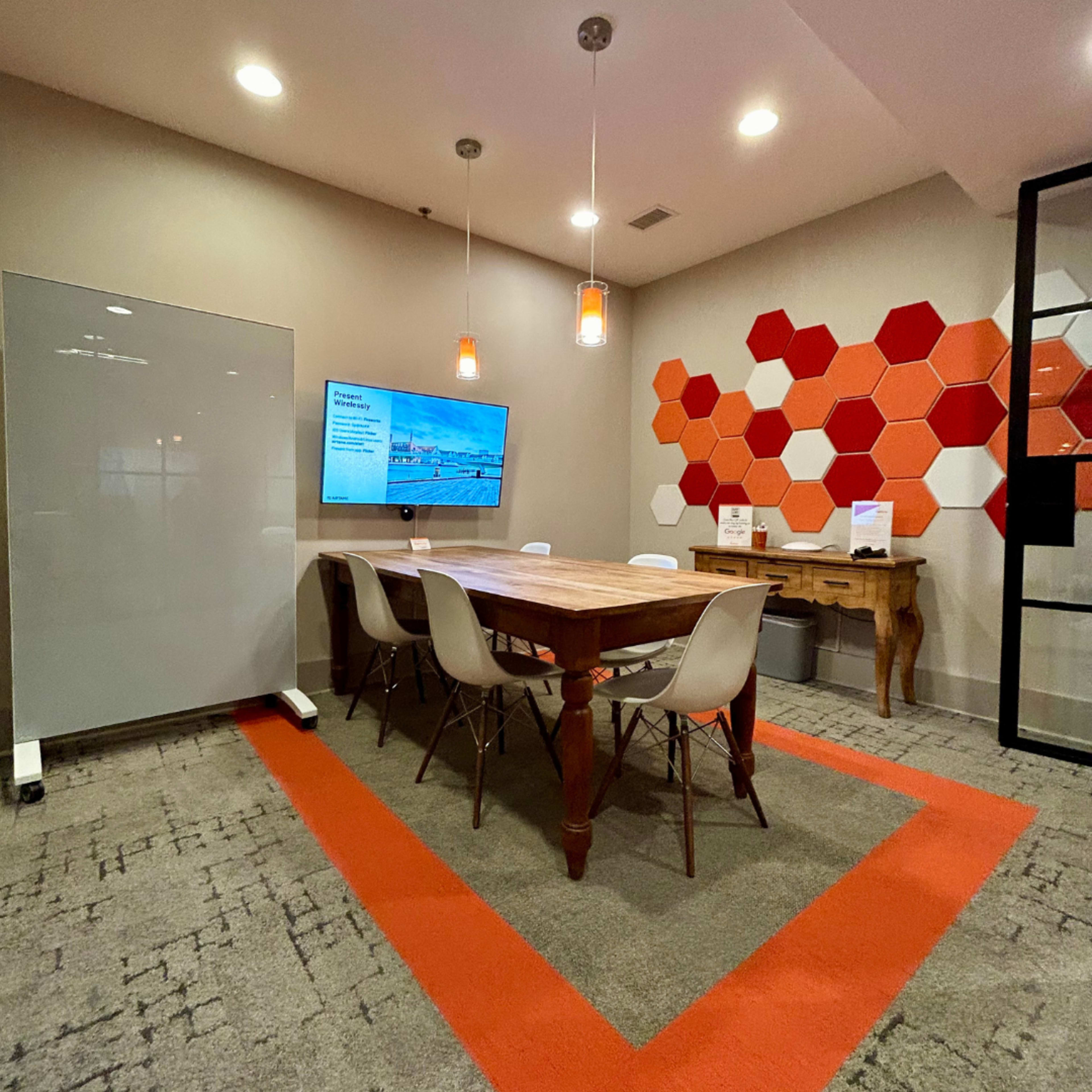 The image shows a modern conference room featuring a wooden table surrounded by white chairs, a wall-mounted screen displaying content, and a colorful hexagonal pattern on one wall.