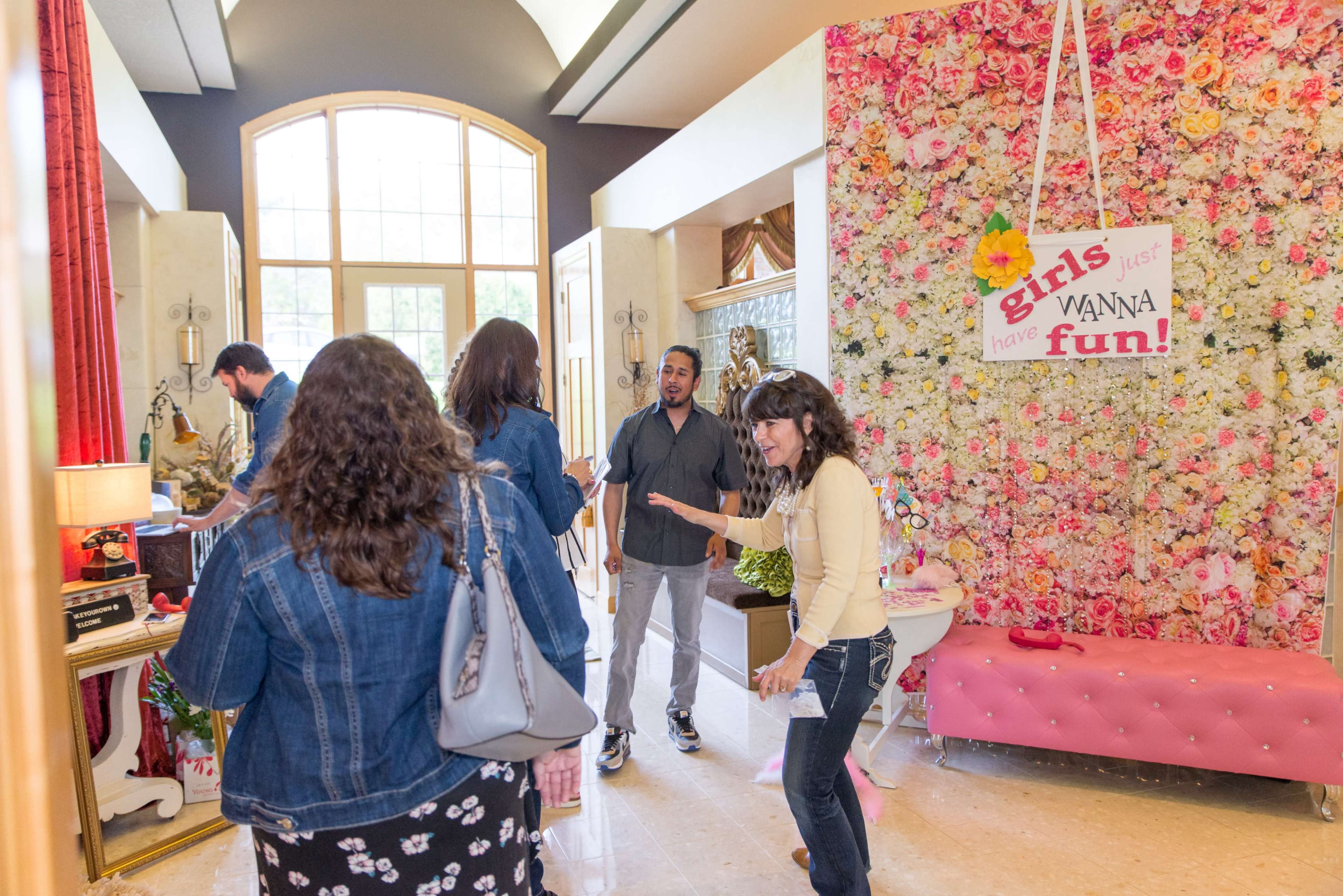 A group of people mingle in a lobby adorned with a vibrant floral backdrop and a sign that reads "girls just wanna have fun!"