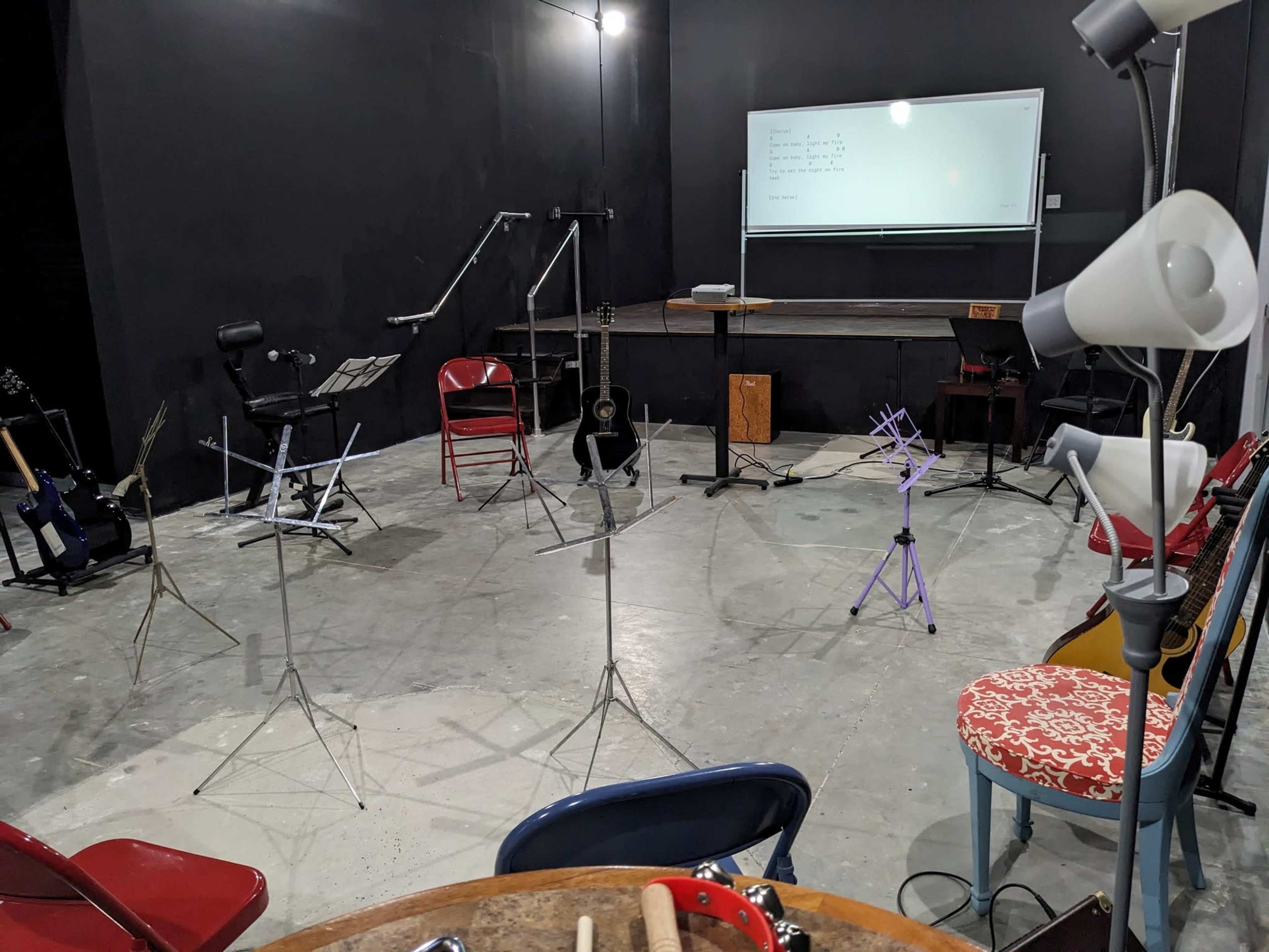 The room features several music stands arranged around a central table, with instruments and chairs positioned near a whiteboard on the wall.