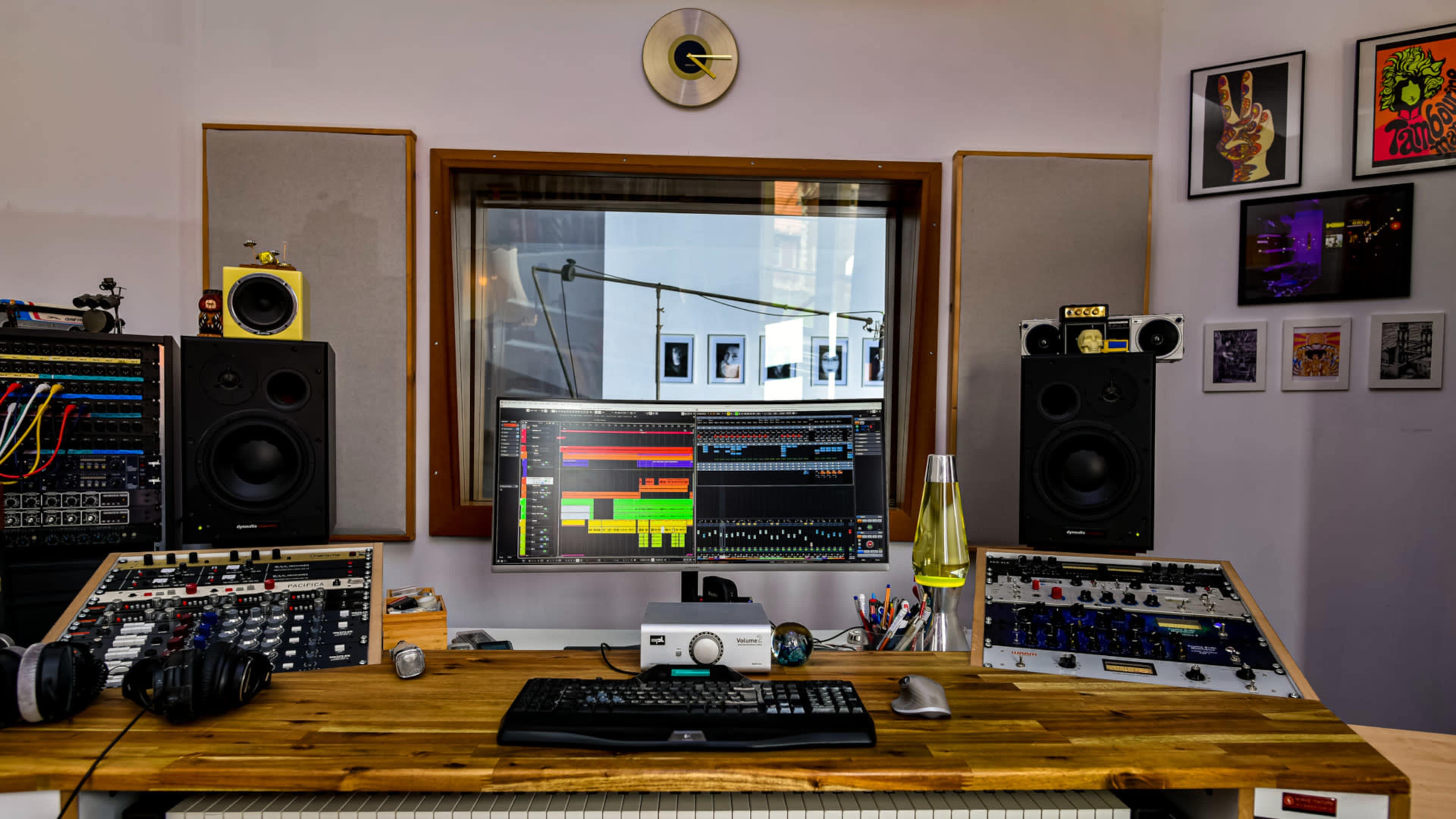 A music production studio with a wooden desk, computer monitors, audio equipment, studio monitors, and framed artwork on the walls.