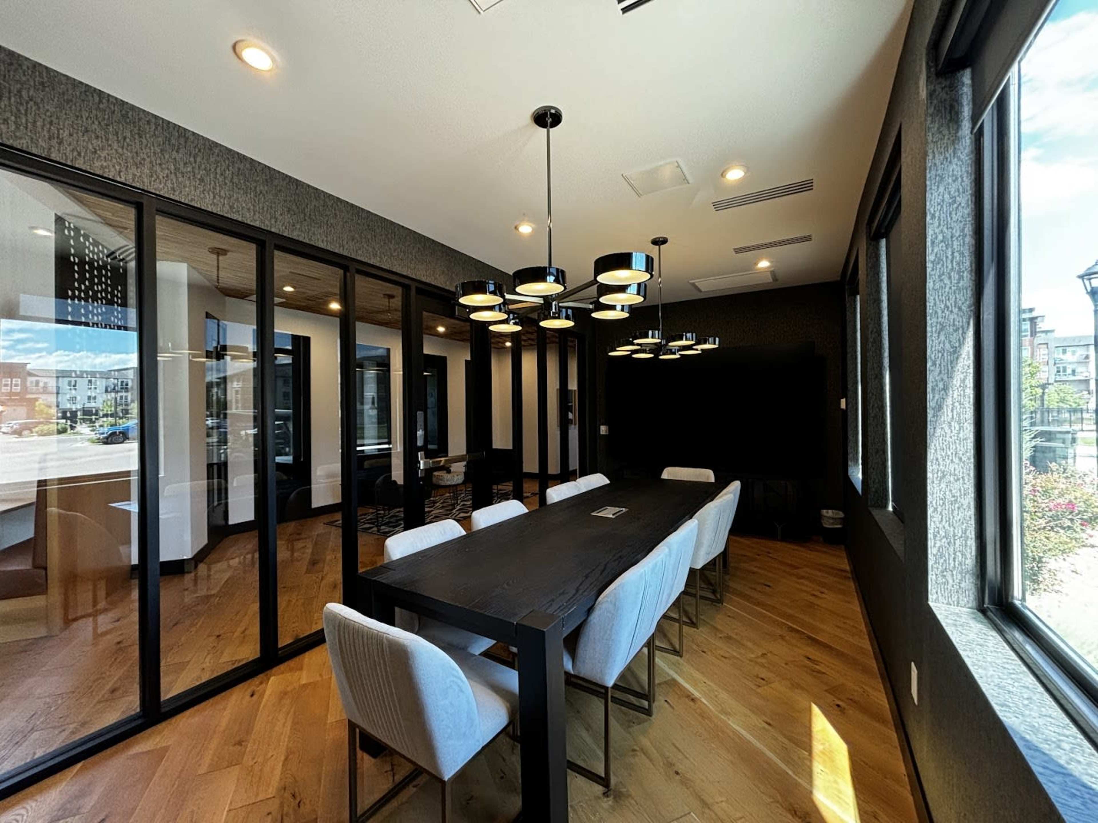A modern conference room features a long wooden table surrounded by upholstered chairs, with large glass panels separating it from the adjoining area.