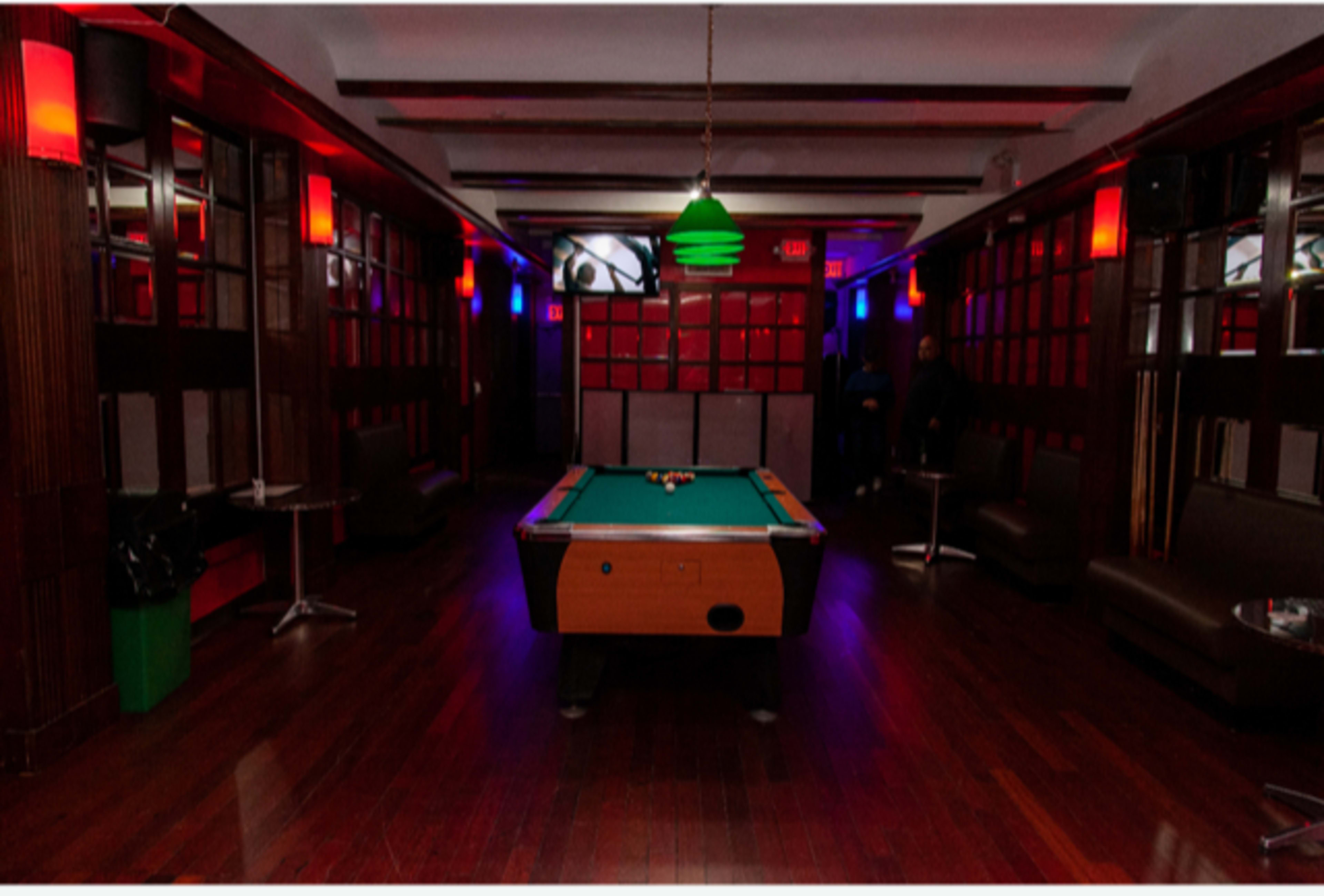 A pool table is positioned in the center of a dimly lit room with red and blue lighting, surrounded by seating areas and a television on the wall.