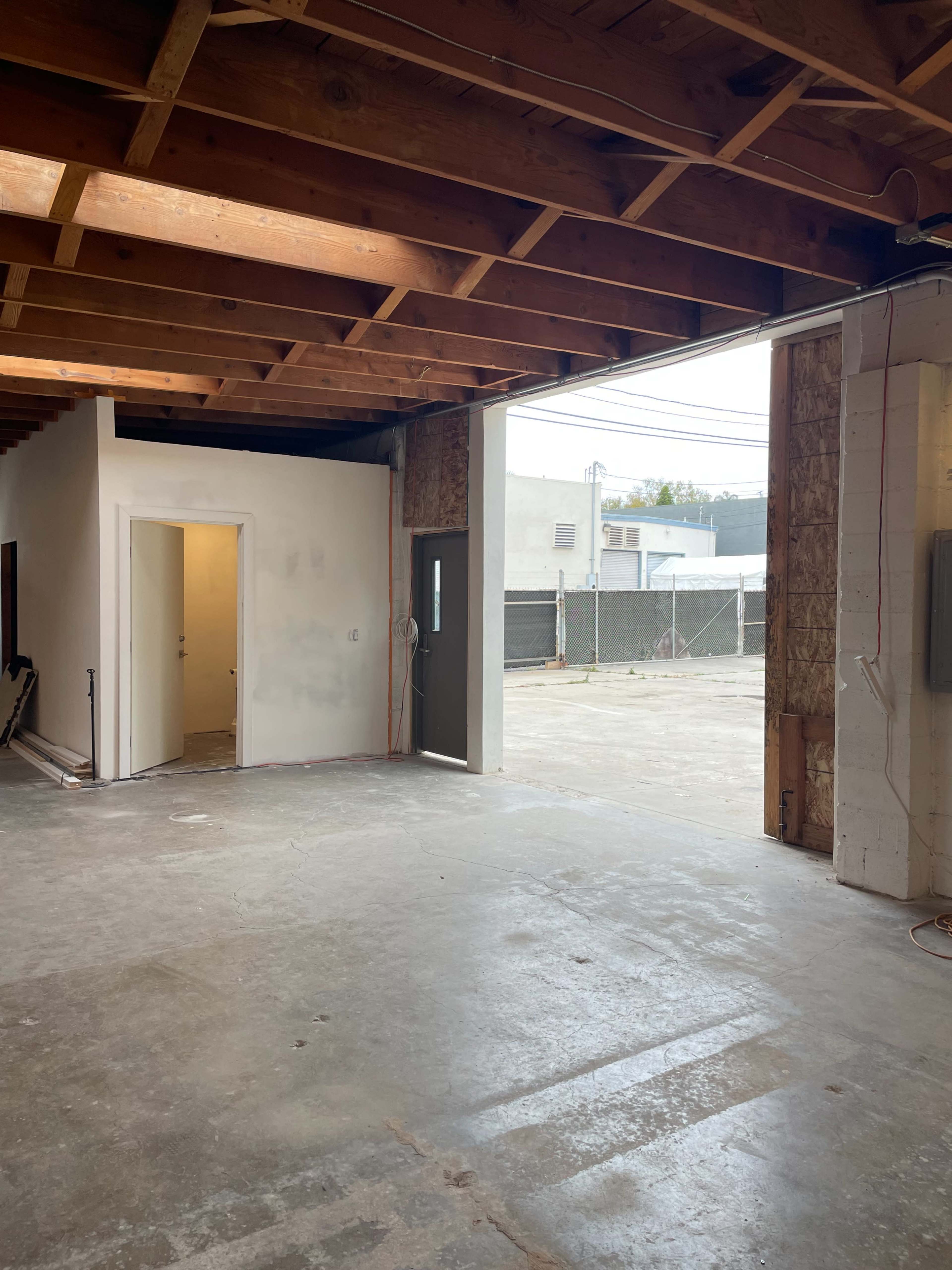 An empty interior space with exposed wooden beams, a doorway leading outside, and a partially finished wall.