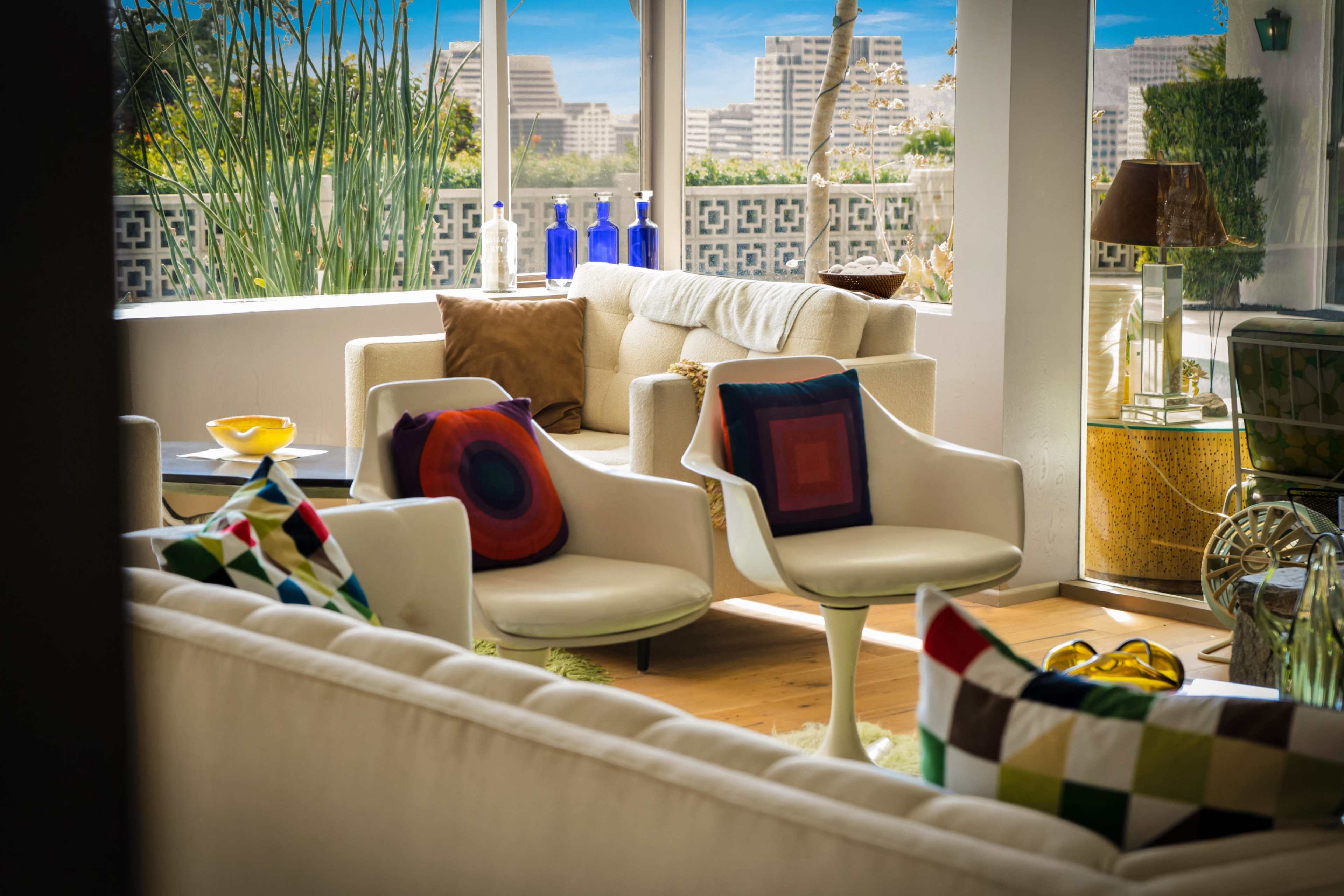 The image shows a modern living room with white furniture, colorful cushions, and large windows overlooking a cityscape.