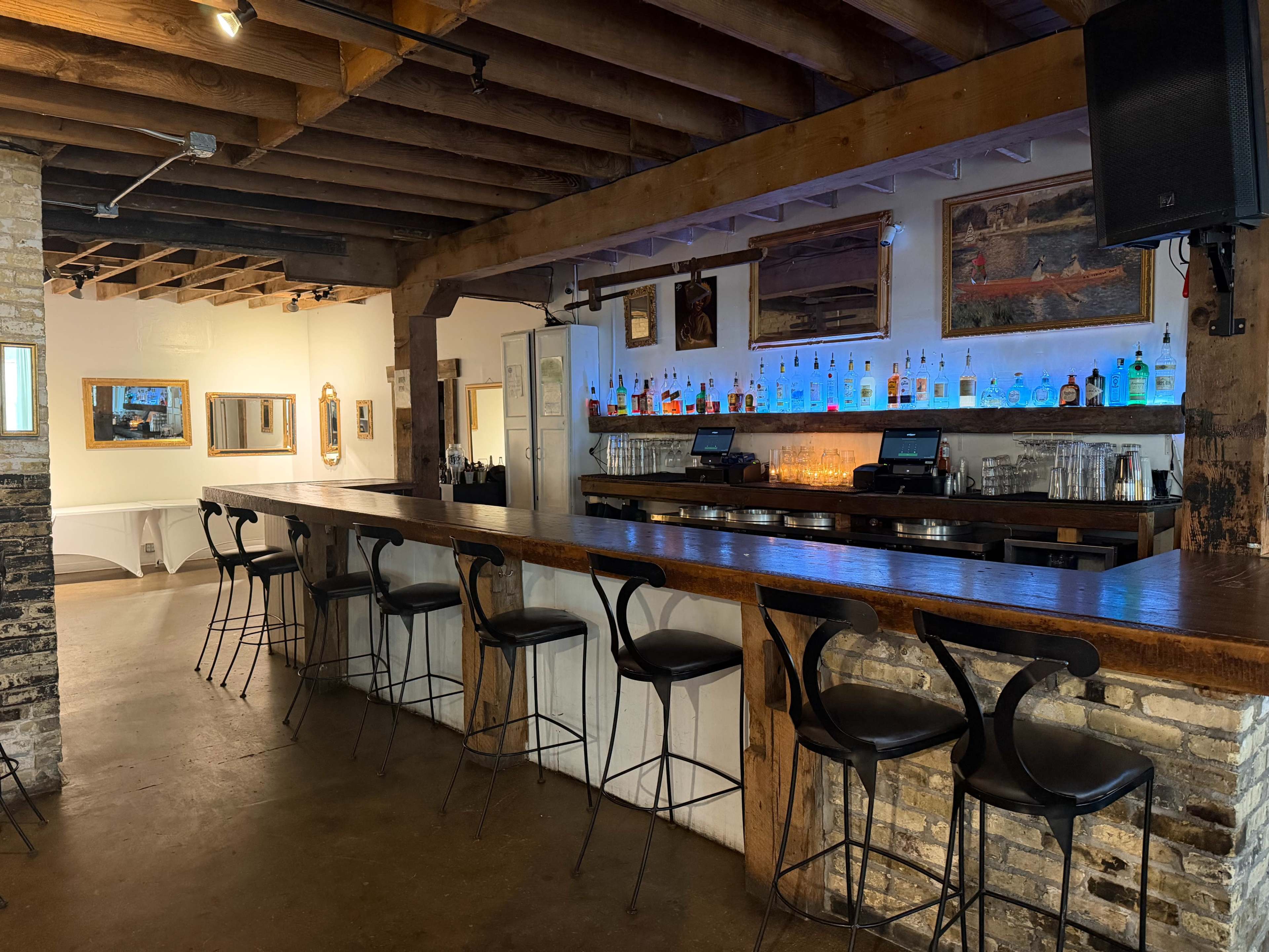 The image shows a well-lit bar area with a long wooden countertop, high stools, and shelves lined with various bottles of liquor set against a rustic backdrop of exposed brick and wooden beams.