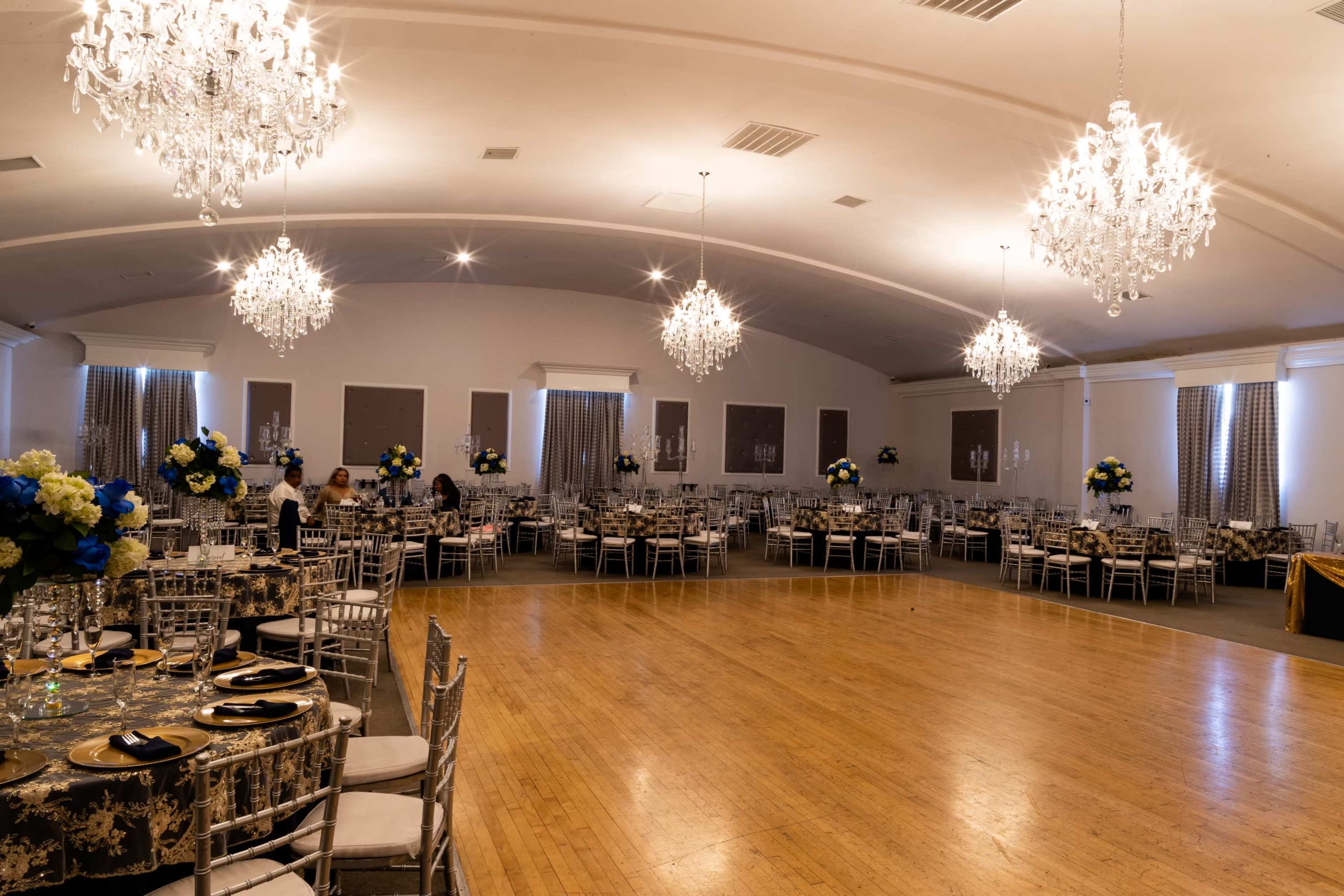 An elegantly decorated banquet hall with a wooden floor, round tables set with floral arrangements, and multiple chandeliers hanging from the ceiling.