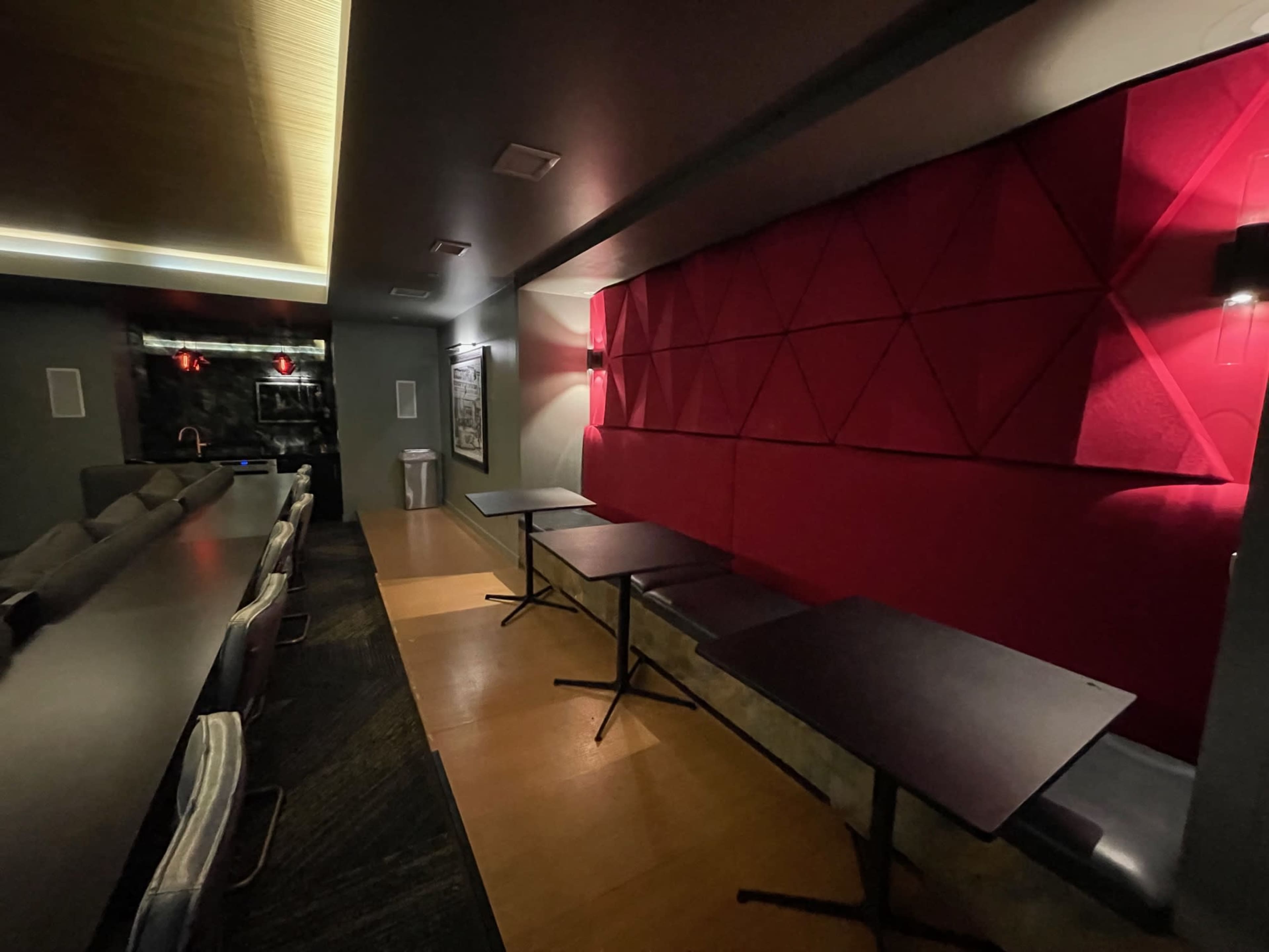 A dimly lit room with a red geometric wall feature, a long table on one side, and several empty tables arranged along the wall.
