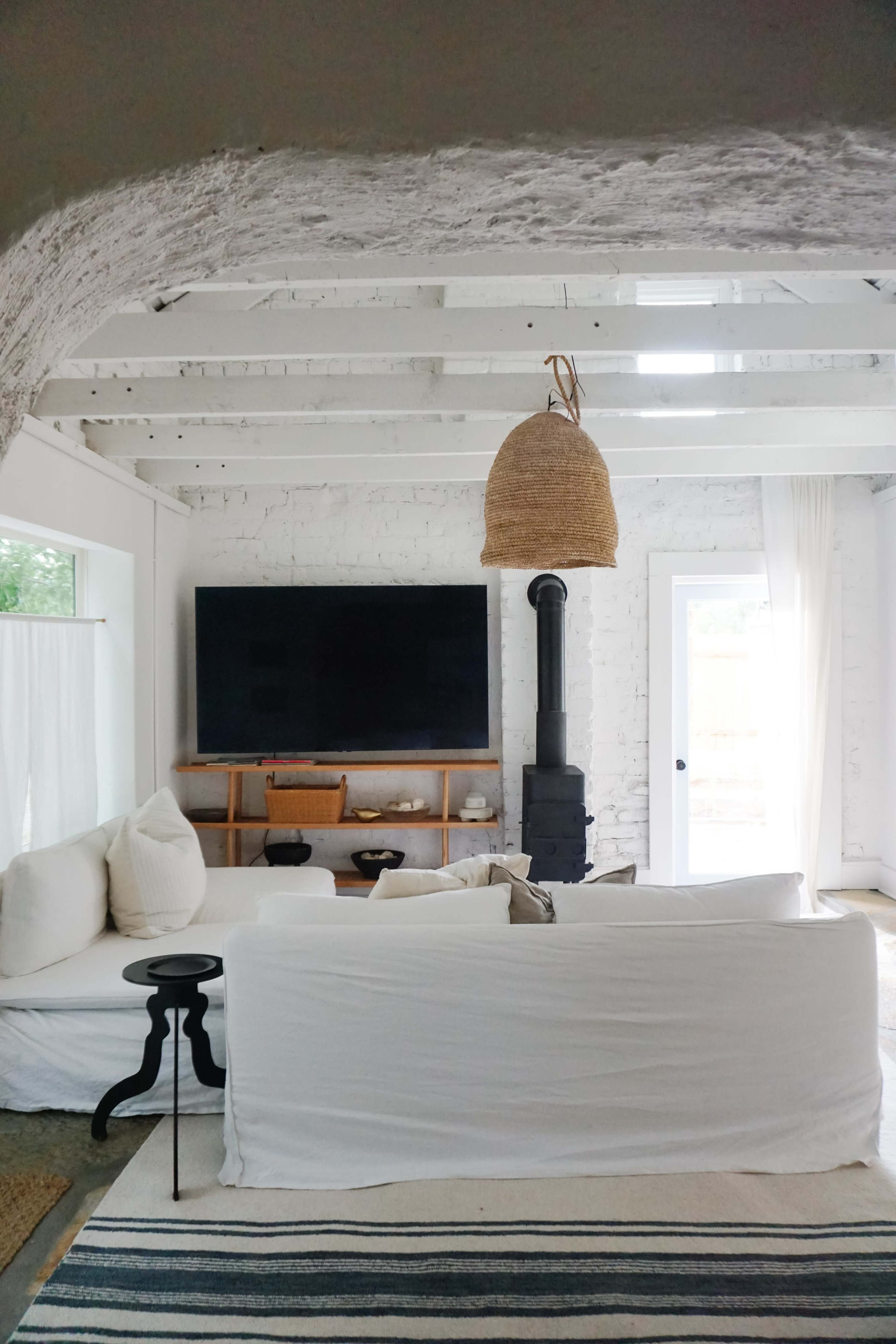 The image shows a cozy living room featuring a white sofa, a black wood stove, a large television mounted on a white brick wall, and a doorway leading outside.