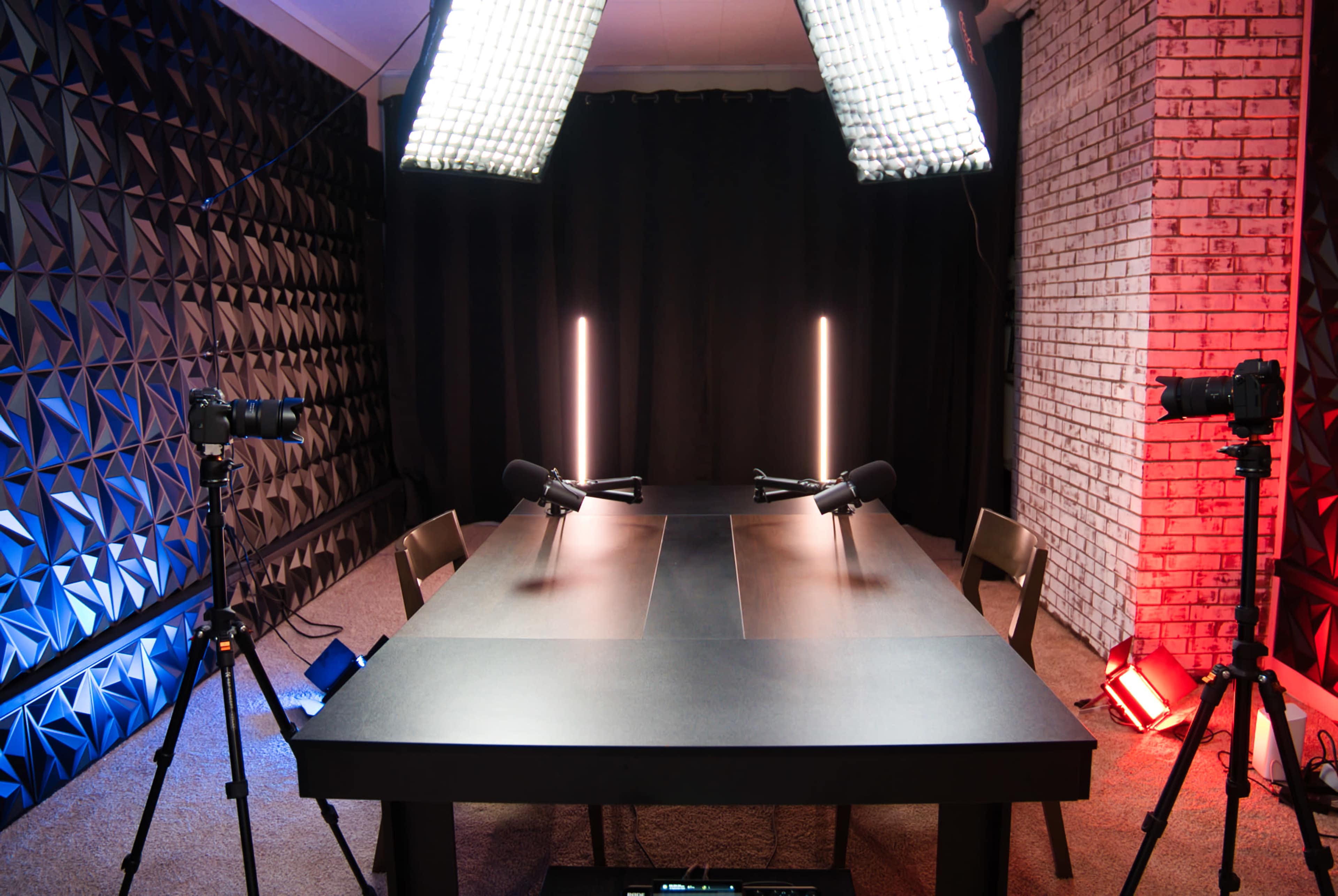 The image shows a podcast recording studio featuring a black table, two microphones, and various lighting setups in a room with textured walls.
