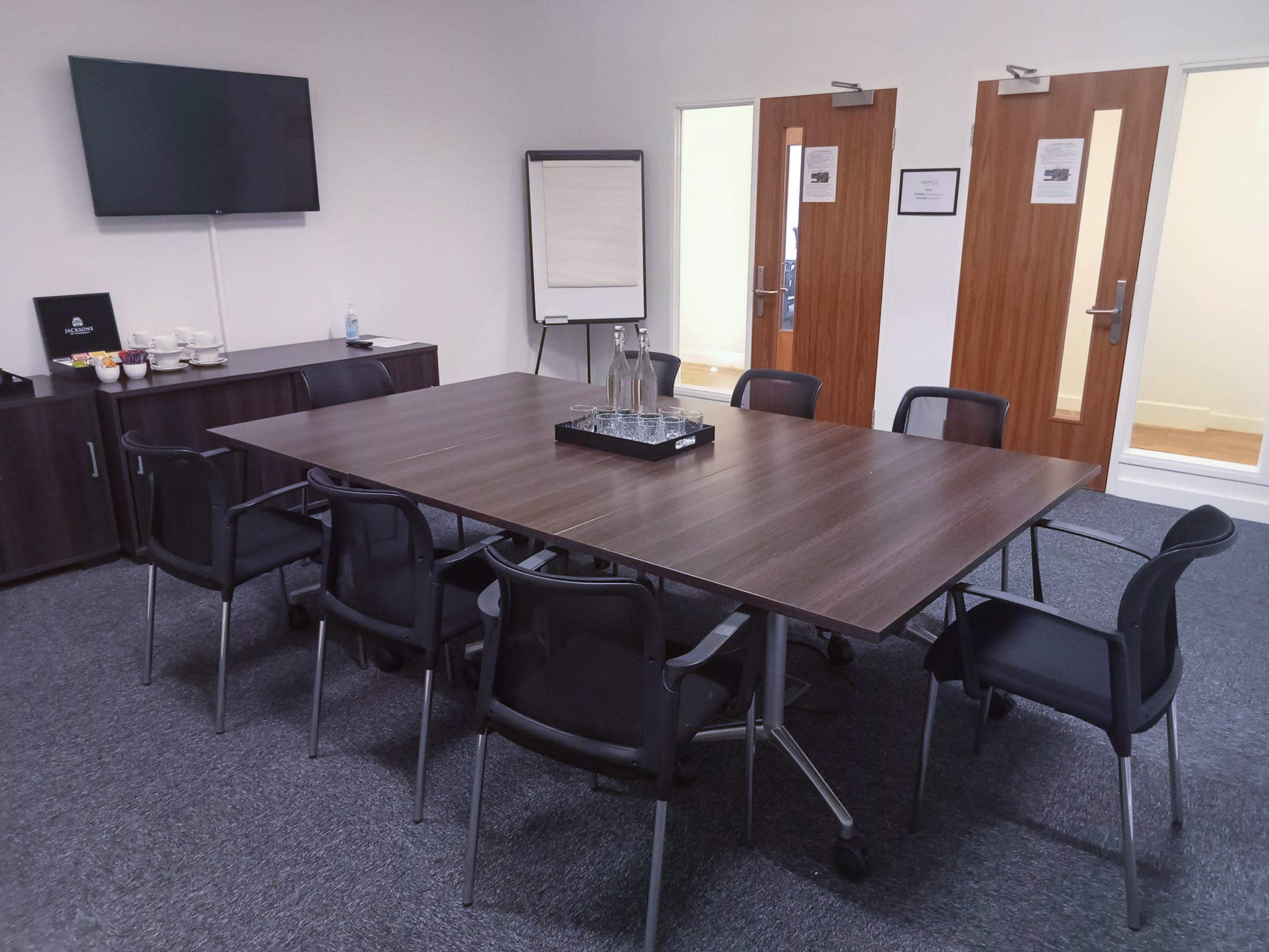 A conference room features a large table surrounded by chairs, with a television and a whiteboard on one wall and two doors on the opposite wall.
