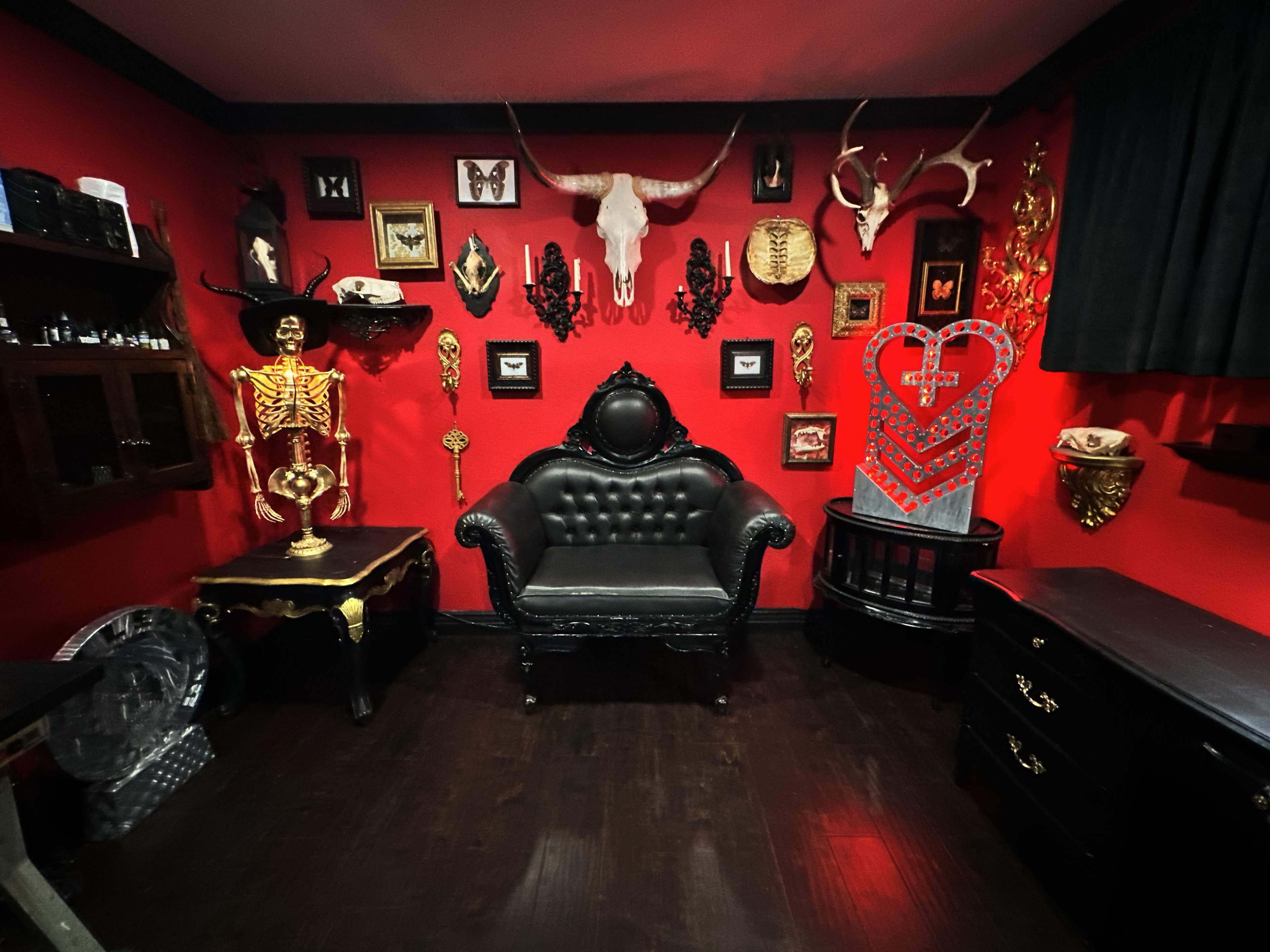 The room features a black vintage chair surrounded by various decorative artifacts, including a skeleton, animal skulls, and framed art on bright red walls.