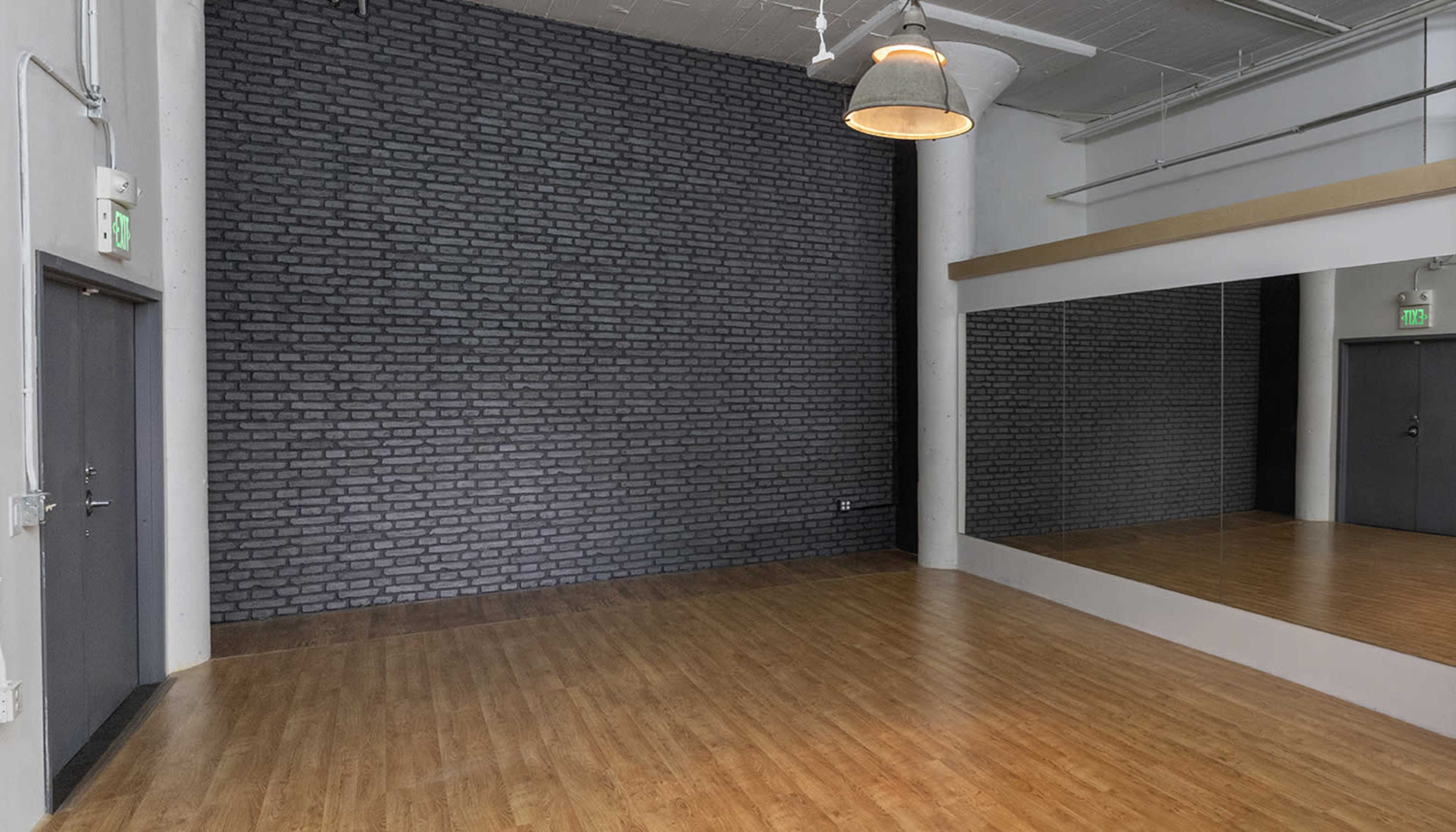 The image shows a spacious room with a wooden floor, a mirrored wall, and a dark brick wall.