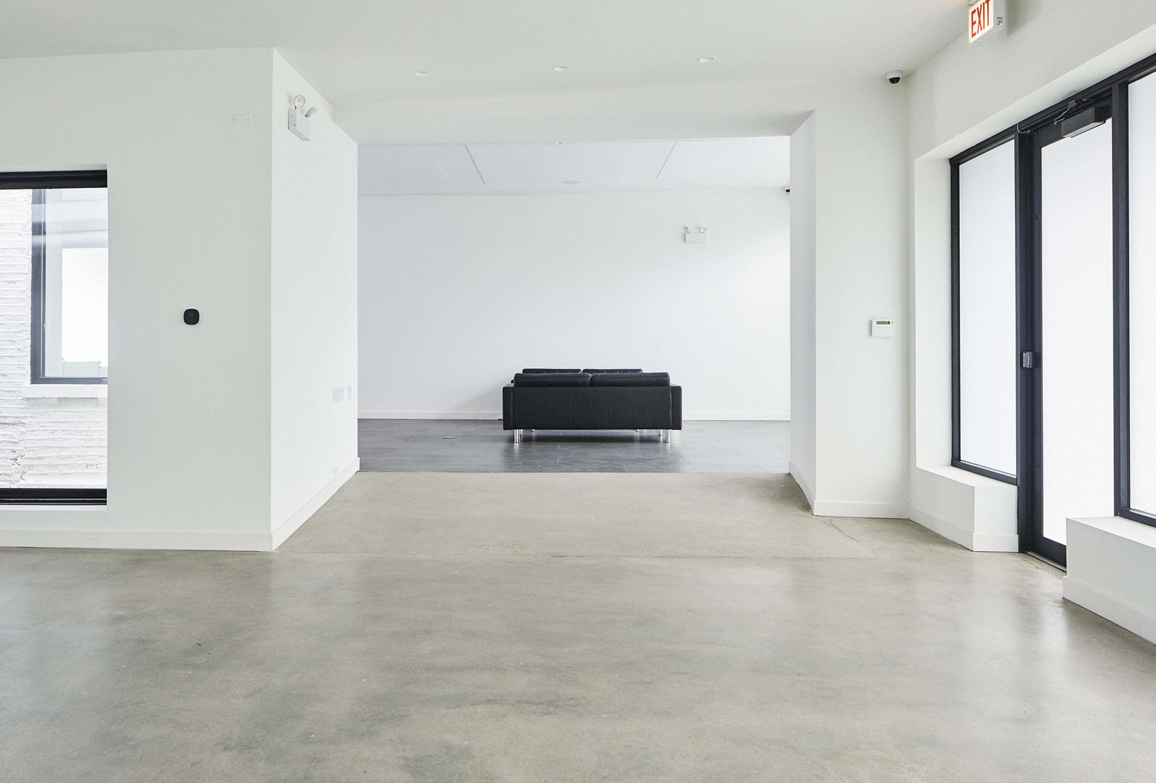 A minimalist interior space with a concrete floor and a black couch in the center, framed by large windows and white walls.