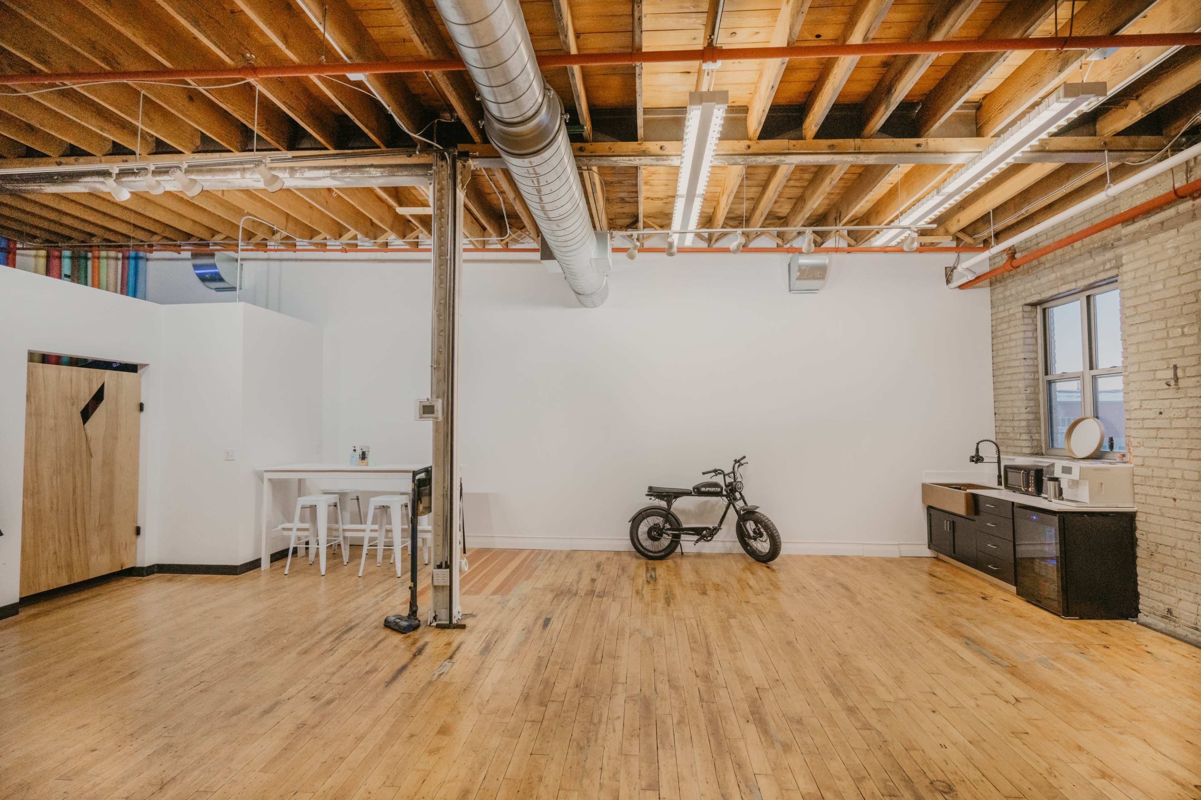 An empty loft space features exposed wooden beams, hardwood floors, a metal support column, and a motorcycle parked against a white wall.