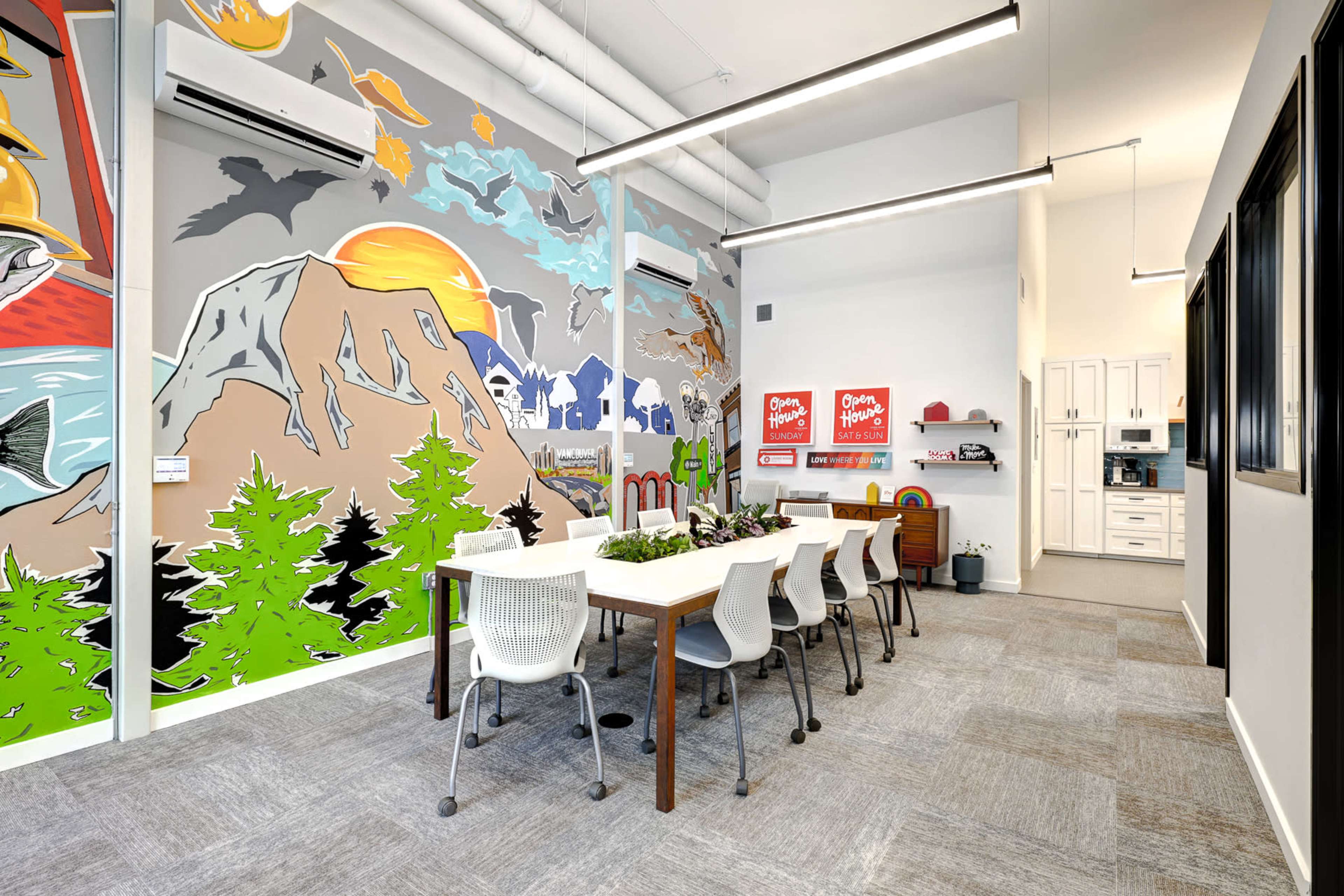 A bright meeting room features a large table surrounded by white chairs, with colorful mural artwork depicting mountains, trees, and wildlife on one wall.