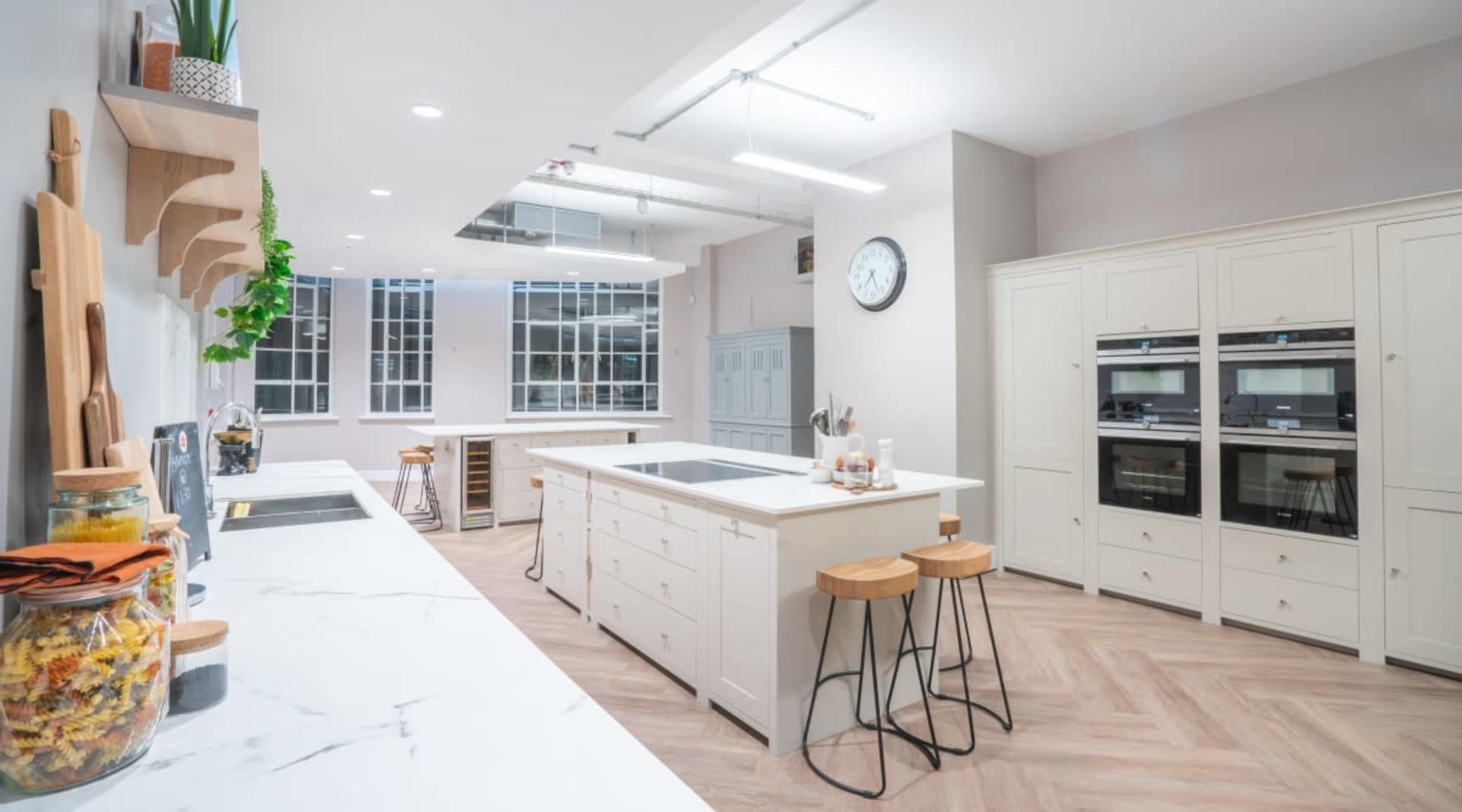 The image shows a modern kitchen featuring a central island with bar stools, built-in ovens, and a large window allowing natural light.