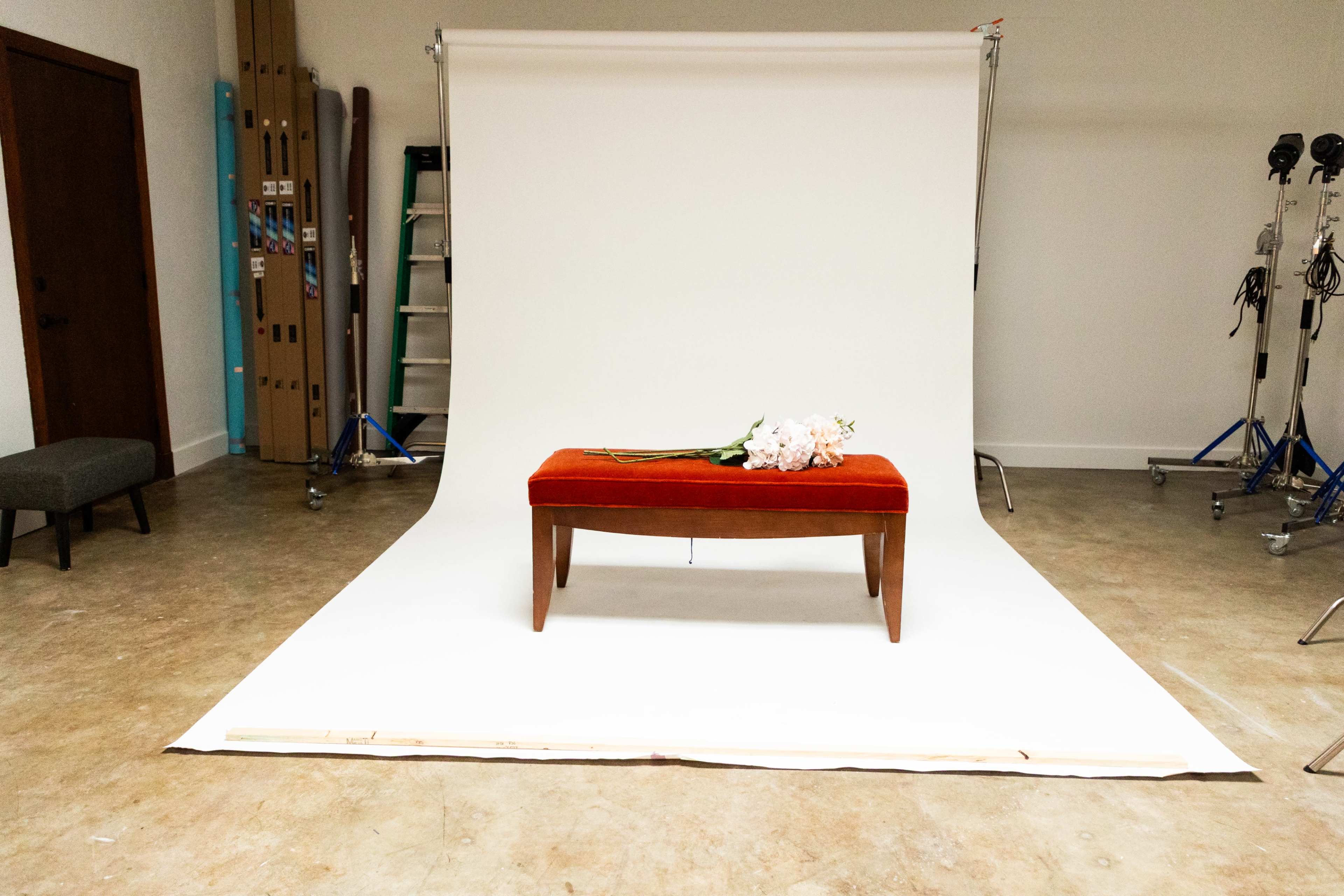 A red upholstered bench with a bouquet of flowers sits on a white backdrop in a photography studio.