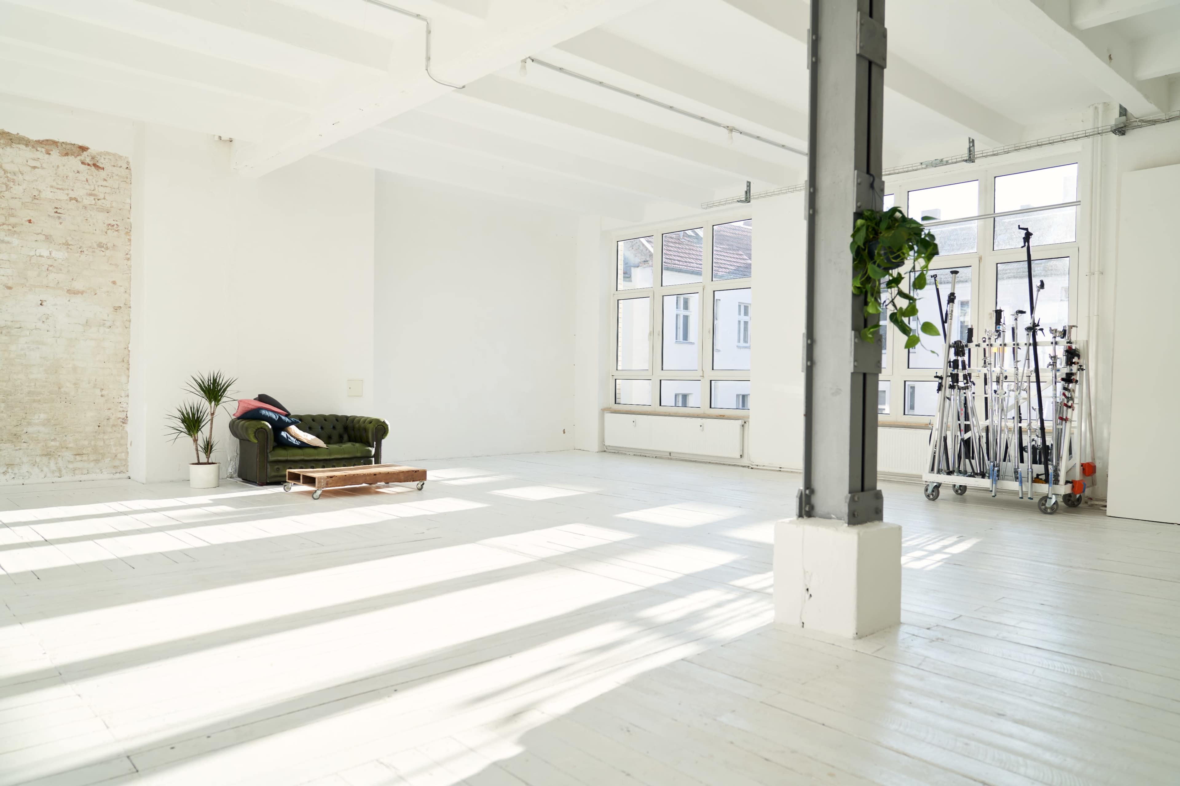 A bright, spacious room with a green couch, a wooden coffee table, and lighting equipment on a cart near the windows.