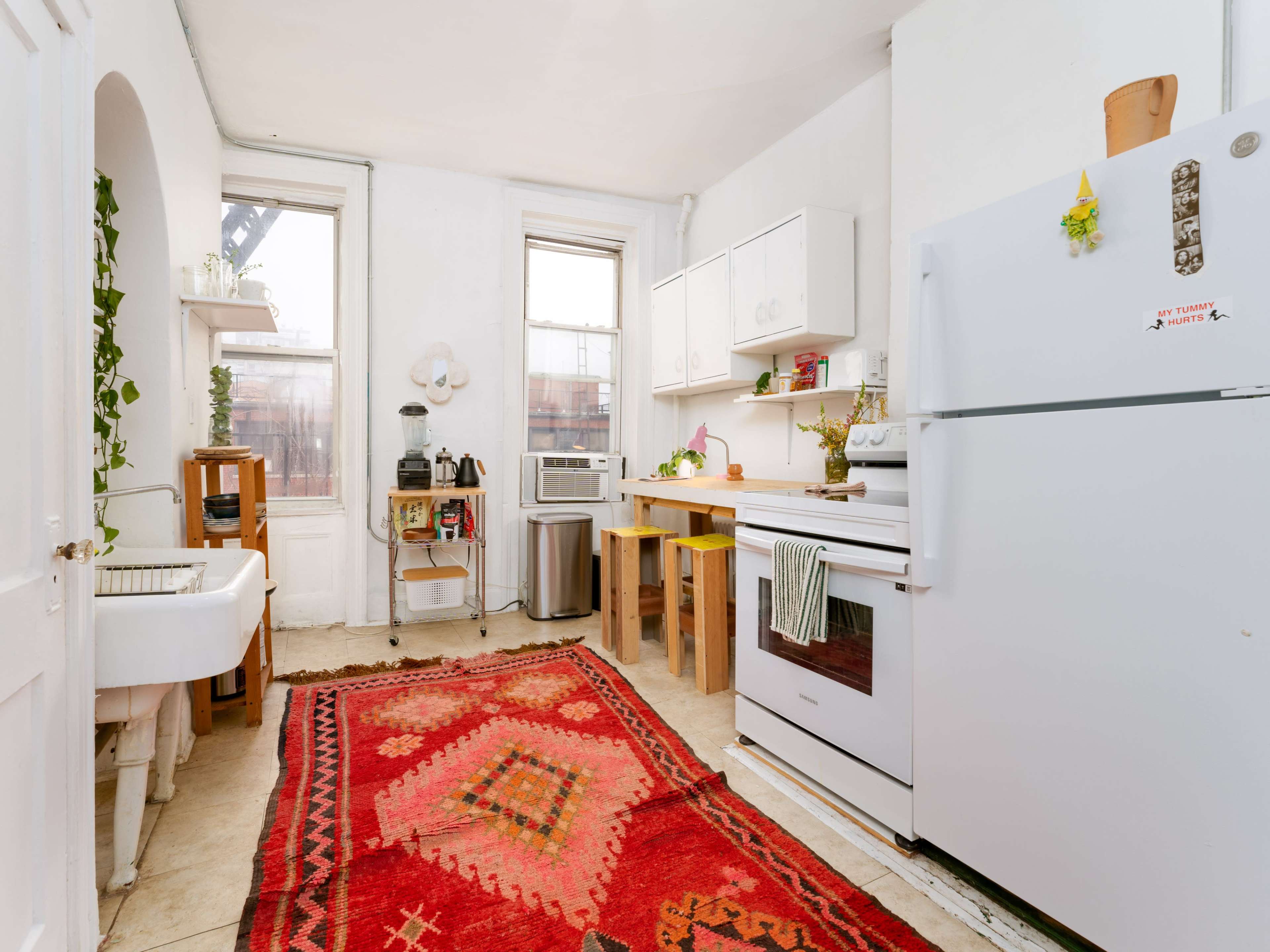 A small kitchen features a white refrigerator, stove, and sink, alongside wooden shelves and a table, with a patterned rug on the floor.