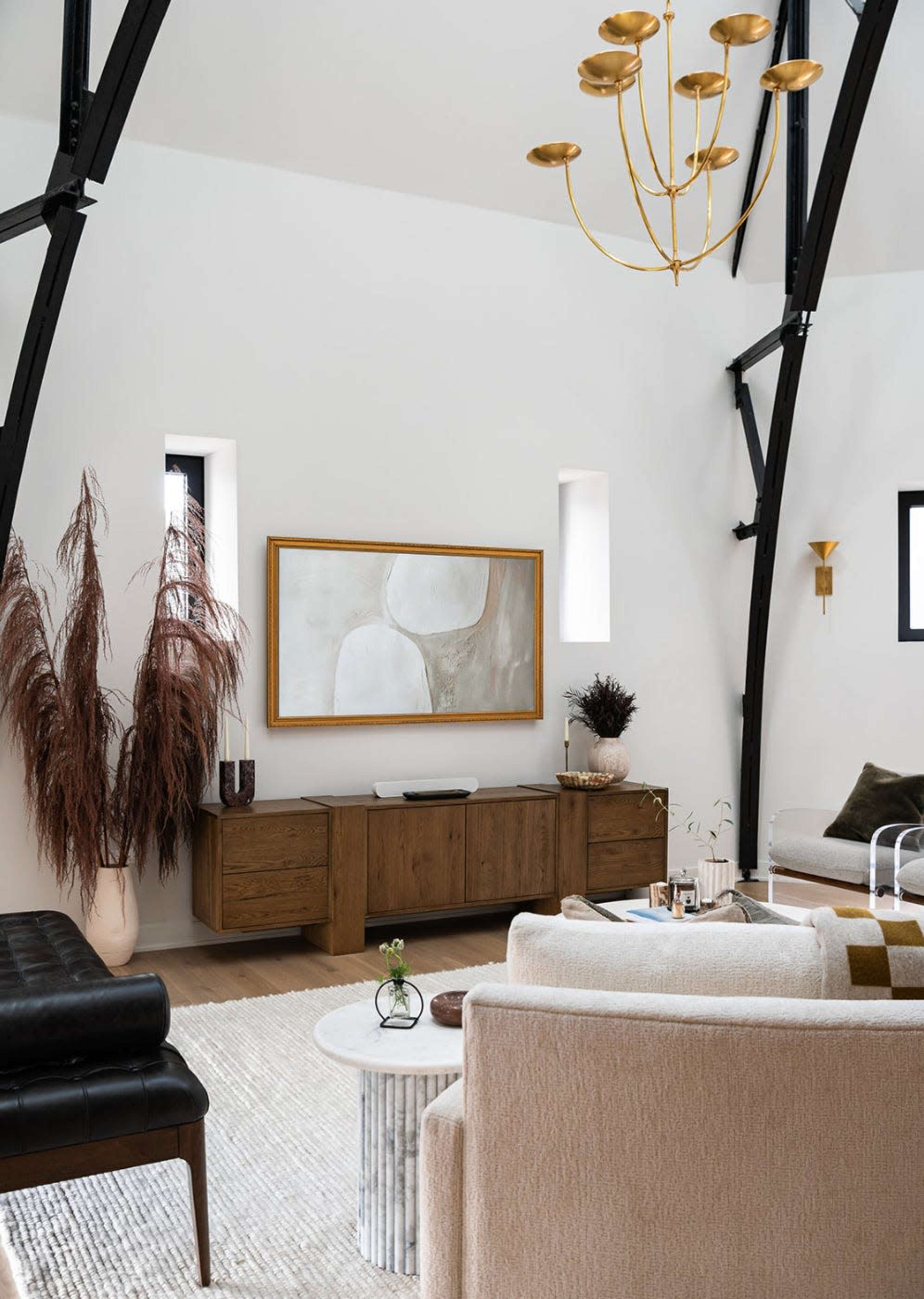 A modern living room features a light color palette, a large abstract painting on the wall, a dark leather sofa, and a mixture of decorative plants and furniture.