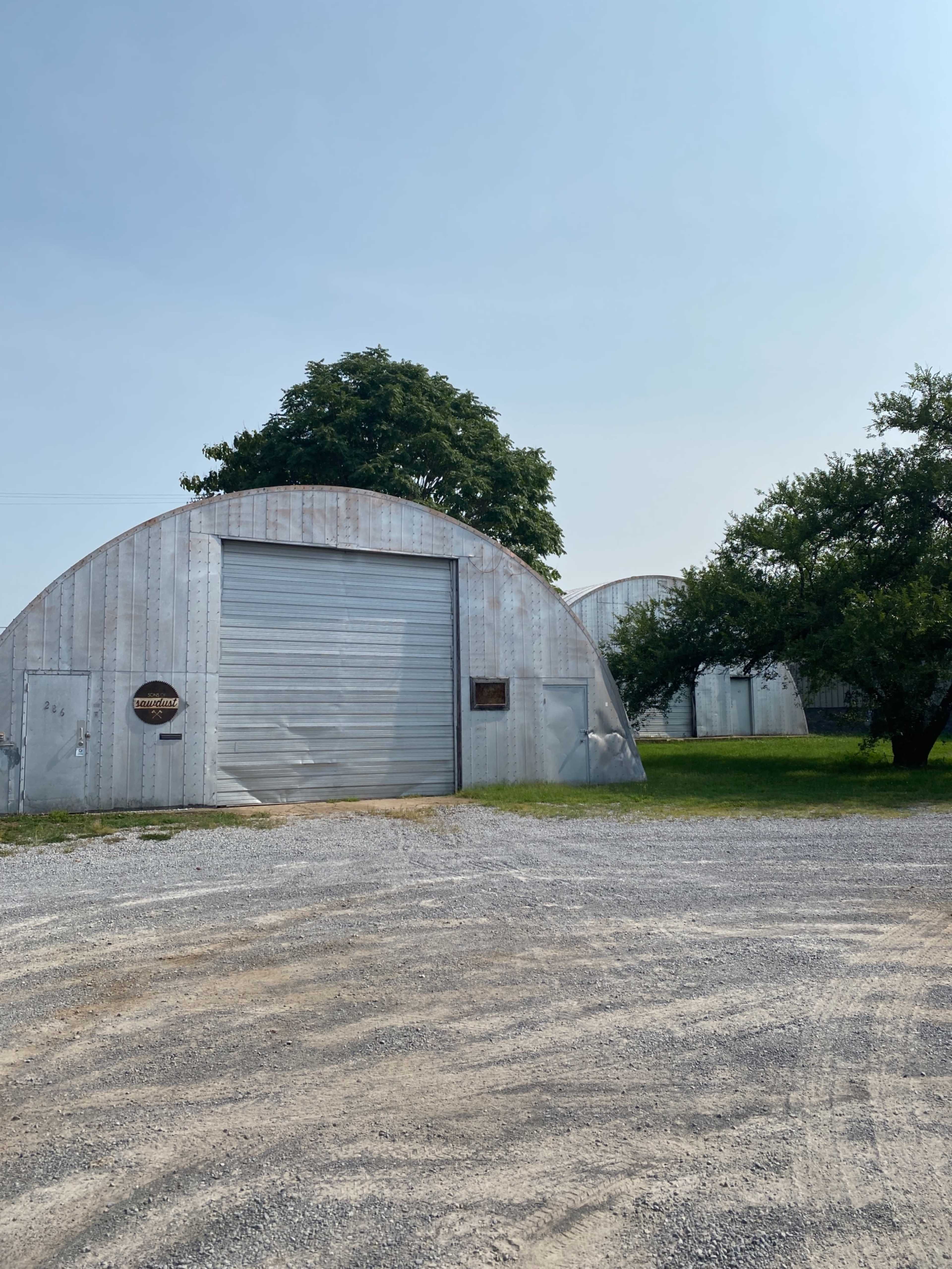 A gravel lot with several large curved metal buildings and a tree nearby.