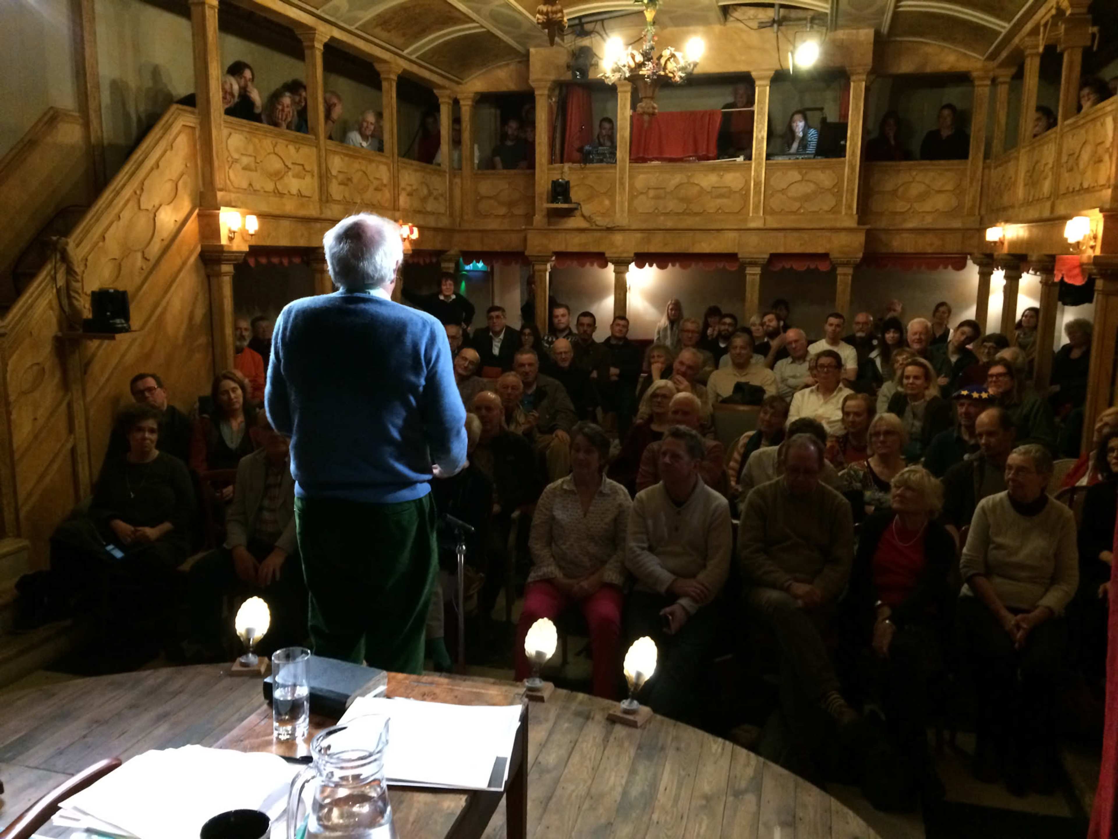A speaker stands on stage addressing a seated audience in a wooden theater filled with spectators.