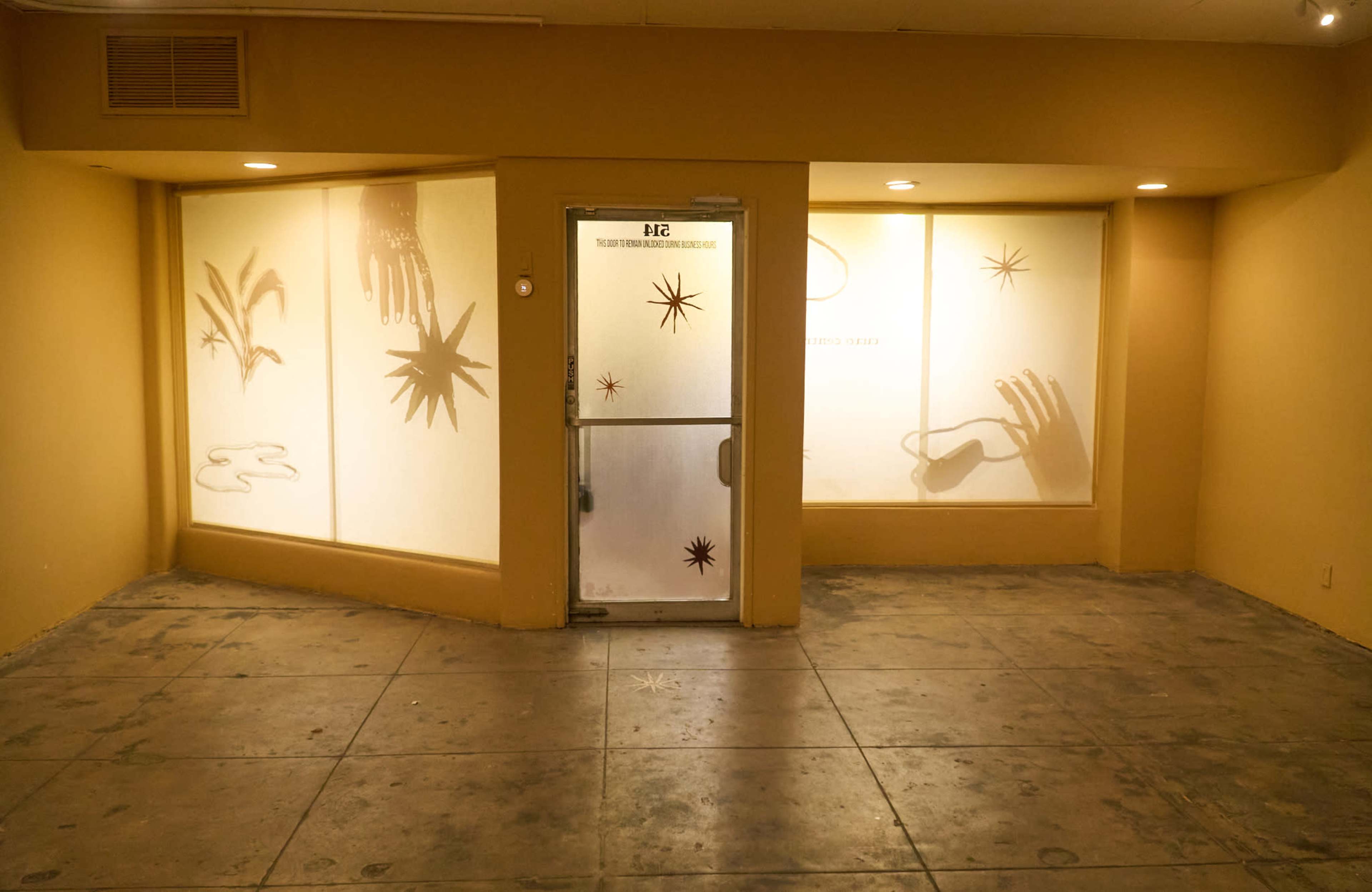 The image shows a vacant interior space with a glass door and large windows displaying artistic silhouettes against a yellow wall.