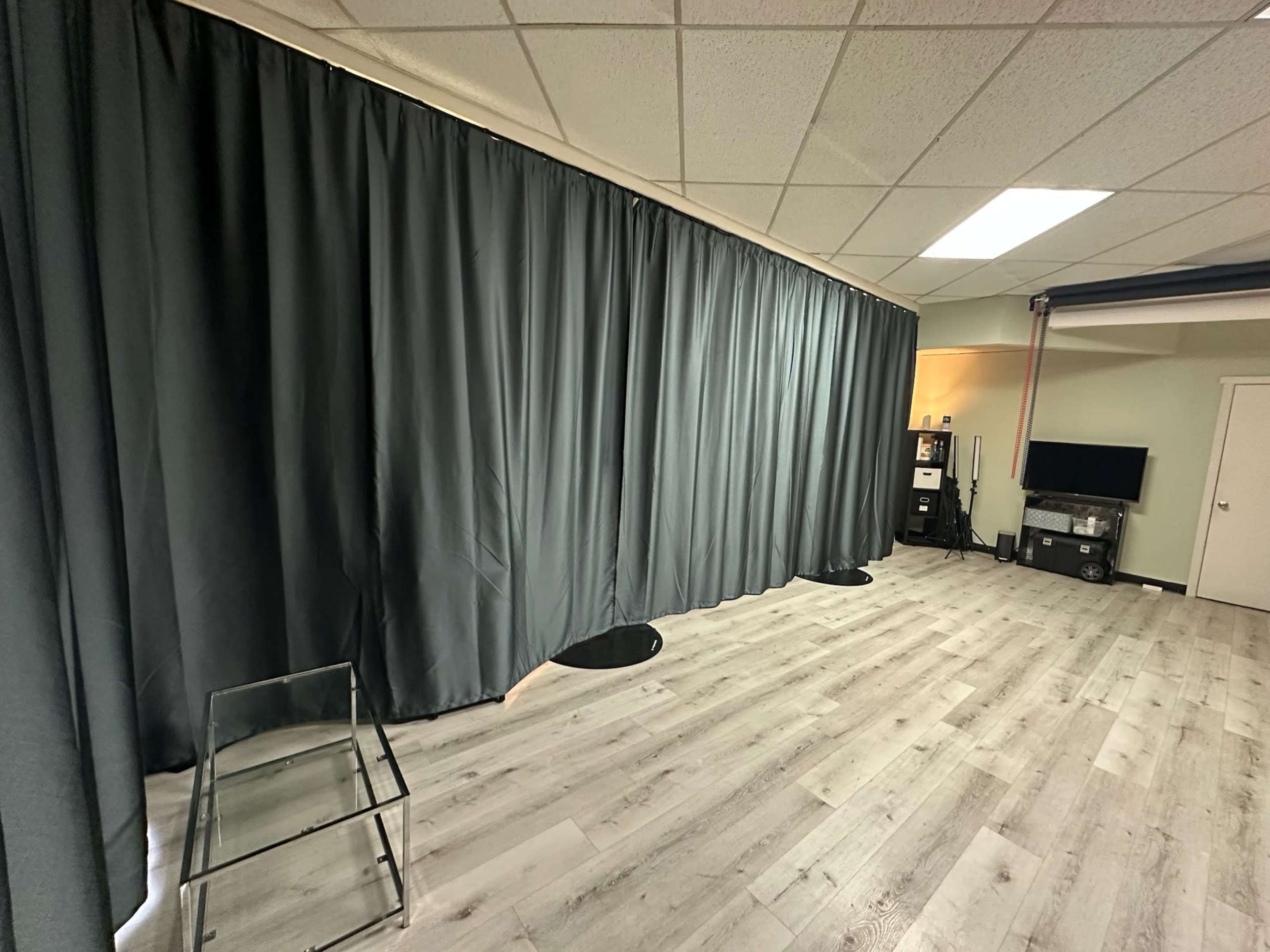 The image shows an interior space with a gray curtain along one side, wooden flooring, and minimal furniture, including a transparent chair and a television setup in the background.