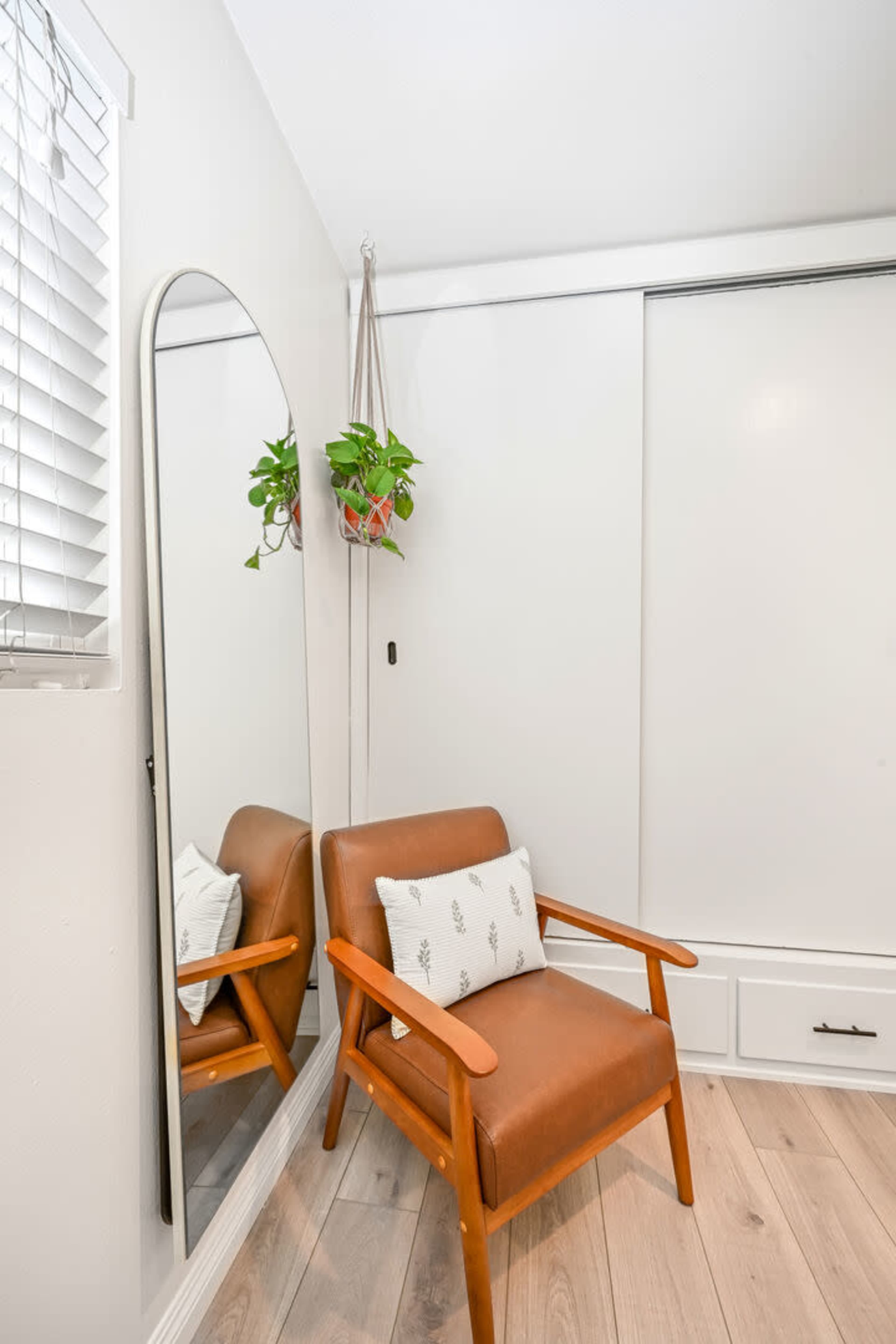 A brown leather chair with wooden arms positioned next to a tall mirror and a hanging potted plant in a bright room with light-colored walls and flooring.