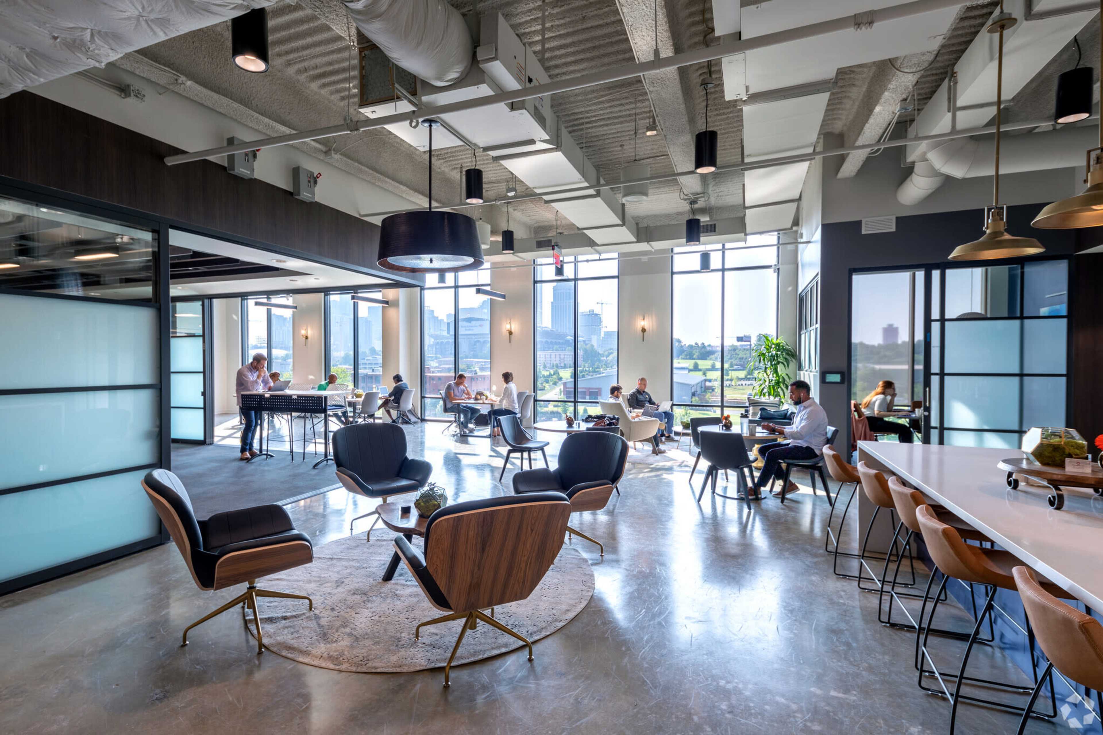 A modern office space with several people working at tables and sitting in lounge chairs, large windows allowing natural light to fill the room.