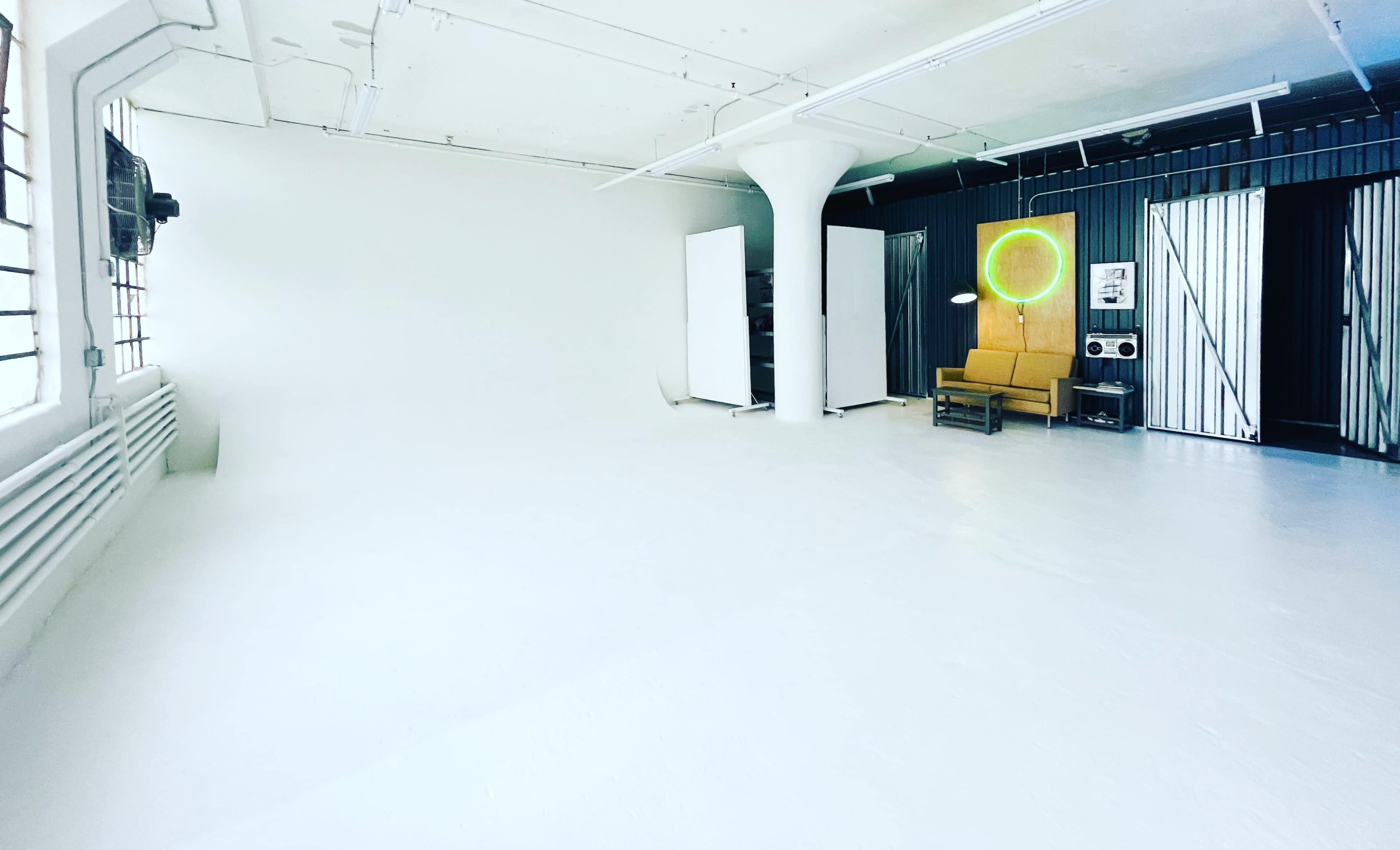 A spacious, well-lit studio with white walls and floor, a yellow couch, and a large circular neon light against a black striped backdrop.
