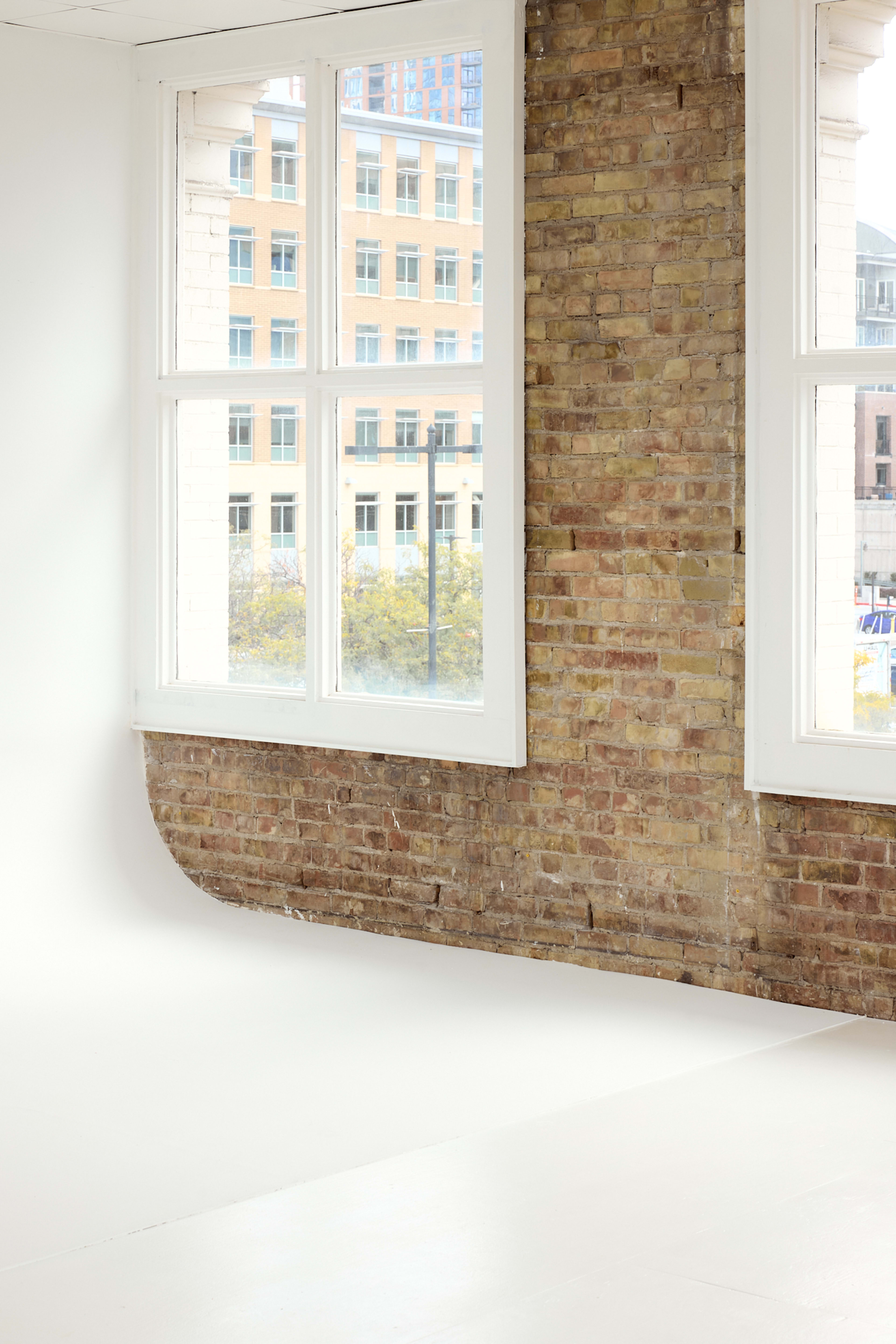 The image shows a bright interior space with large windows, featuring exposed brick walls and a smooth, white floor.